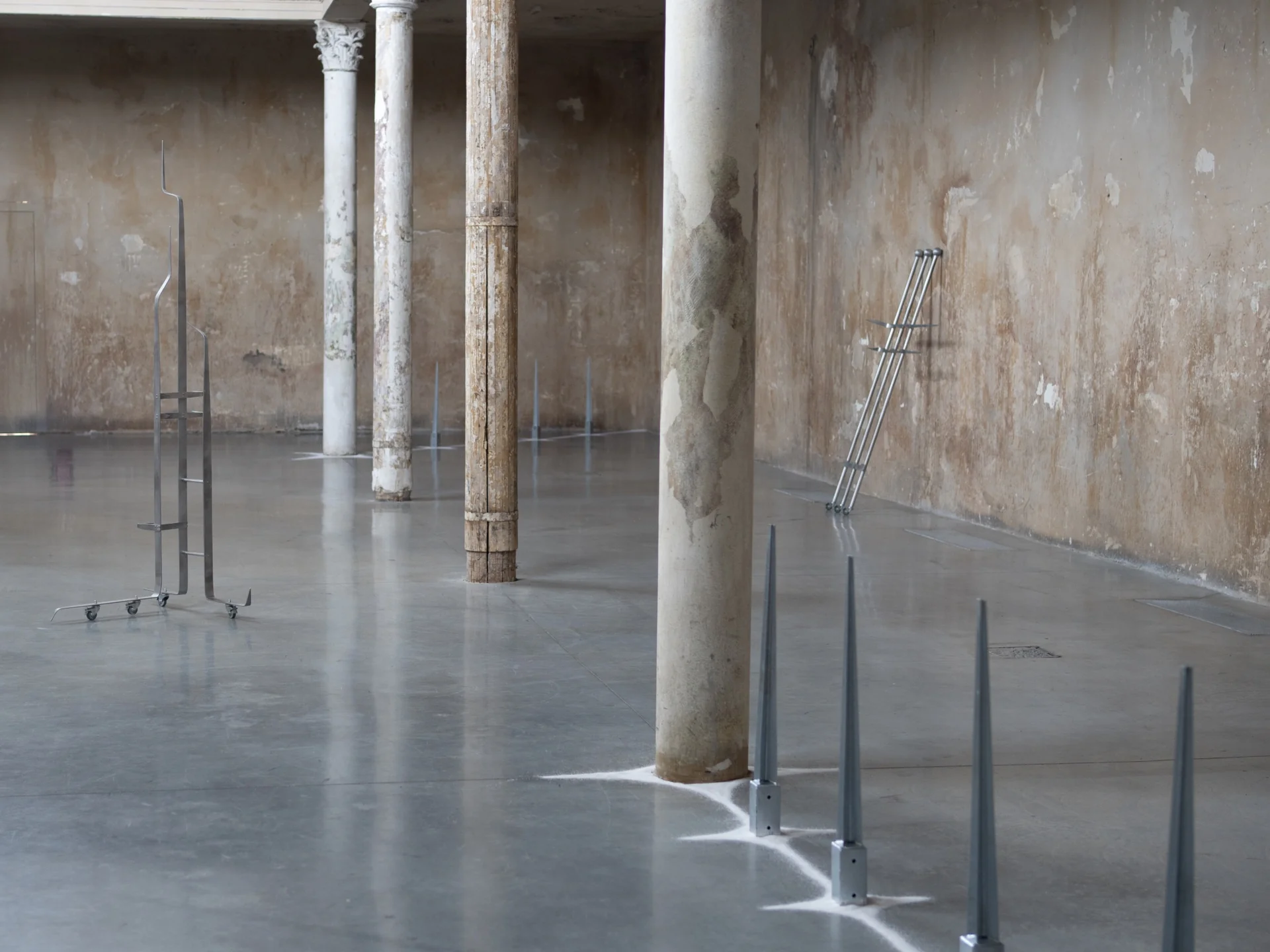 Everything Comes Together While Pushing All Apart by Roxana Morar — Galvanized spike stakes by Albert Kaan rising from polished concrete next to Andrei Arion's wheeled stainless ladder, Corinthian column shedding plaster between them.