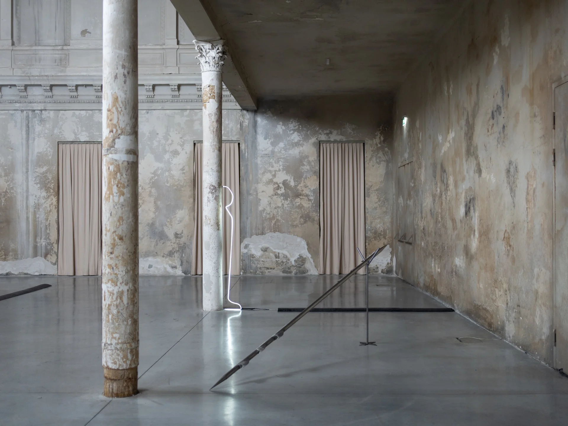 Everything Comes Together While Pushing All Apart by Roxana Morar — Neon outline of a standing figure by Ana Ionescu lit against an interior column, two arrow-tipped steel rods by Albert Kaan grounded in the foreground floor.
