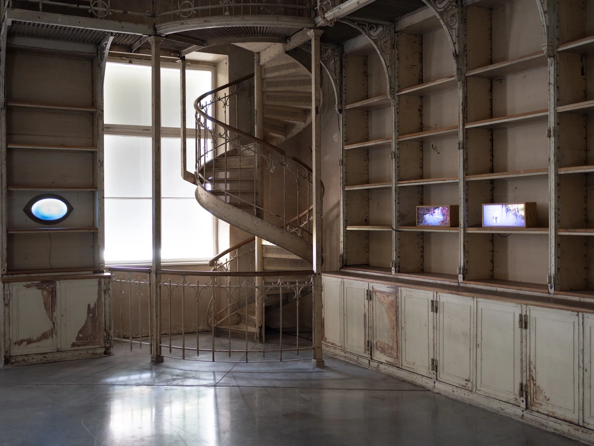 Everything Comes Together While Pushing All Apart by Roxana Morar — Spiral cast-iron staircase and emptied library cabinetry, two small monitors by Albert Kaan resting on a shelf with an oval portal cut into the joinery below.