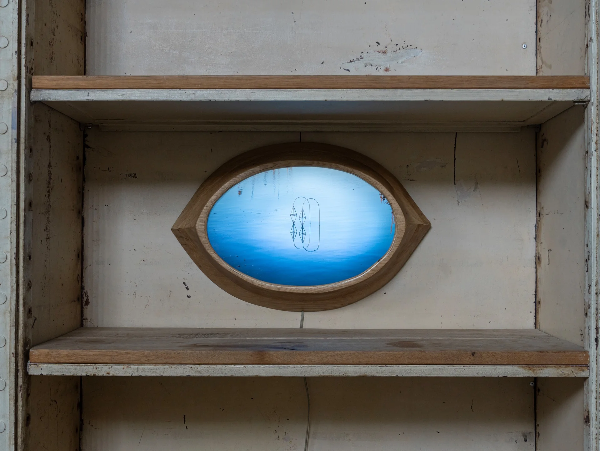 Everything Comes Together While Pushing All Apart by Roxana Morar — Eye-shaped oak frame by Albert Kaan housing a small monitor of blue water and a tiny boat, set inside a stripped wooden bookcase.