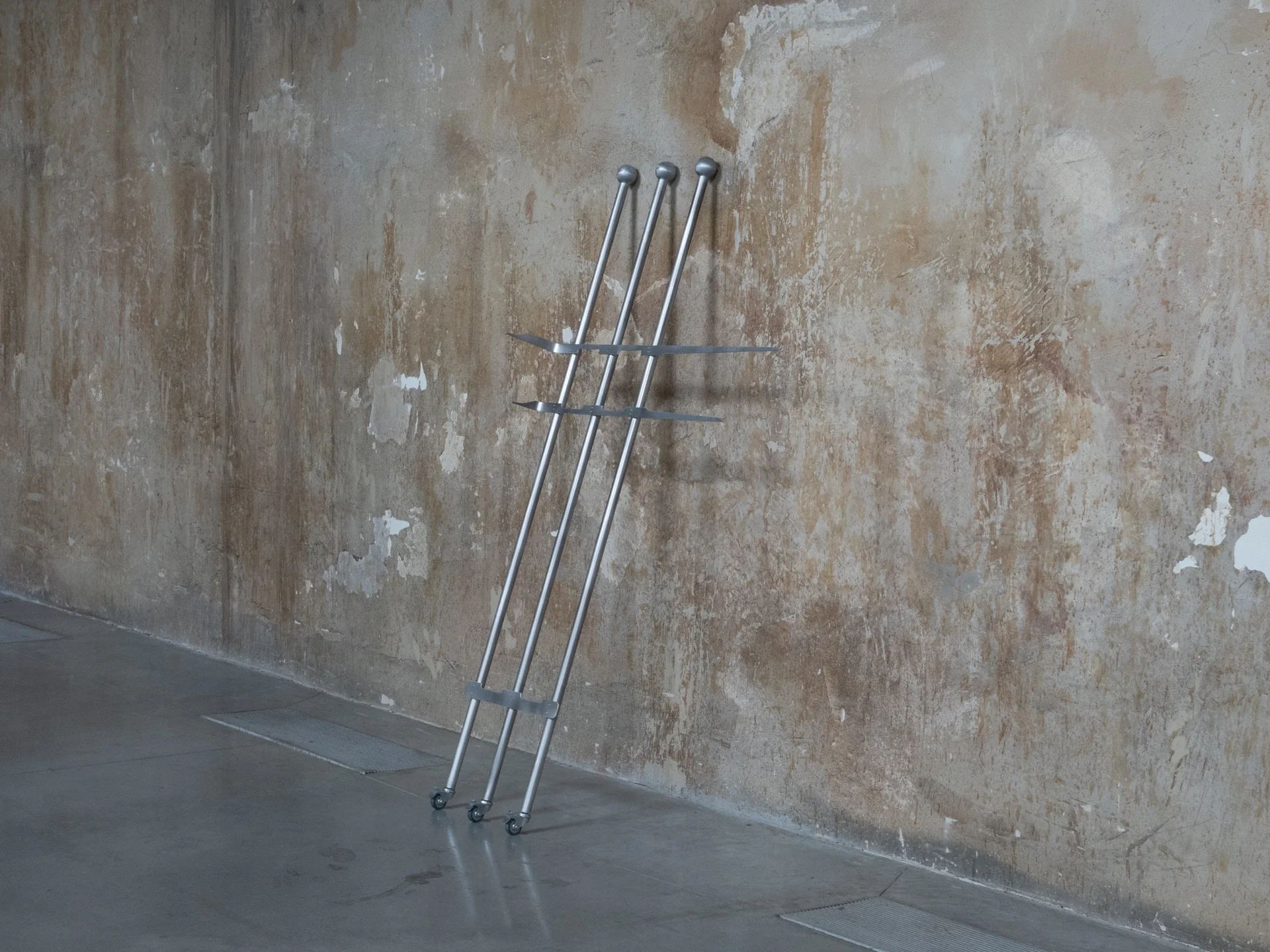 Everything Comes Together While Pushing All Apart by Roxana Morar — Three stainless ball-tipped poles by Andrei Arion leaning together against the peeled-back paint of the gallery wall, small casters at their feet.