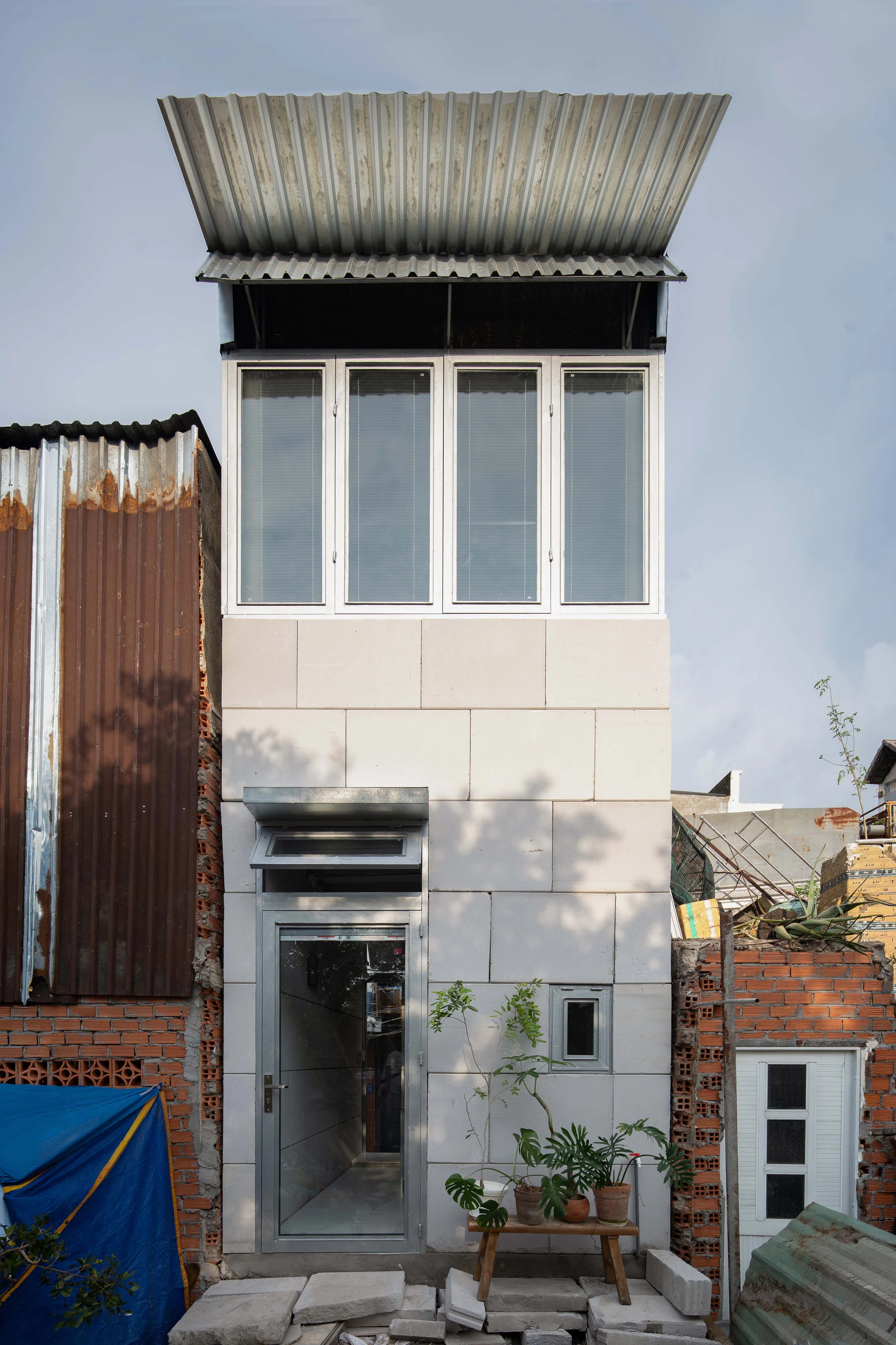 11M2 HOUSE by scale50, photographed by Nguyen Manh Toan — Upper room at daylight, a figure adjusts the blinds against a ribbon of casement windows, OSB ceiling overhead.