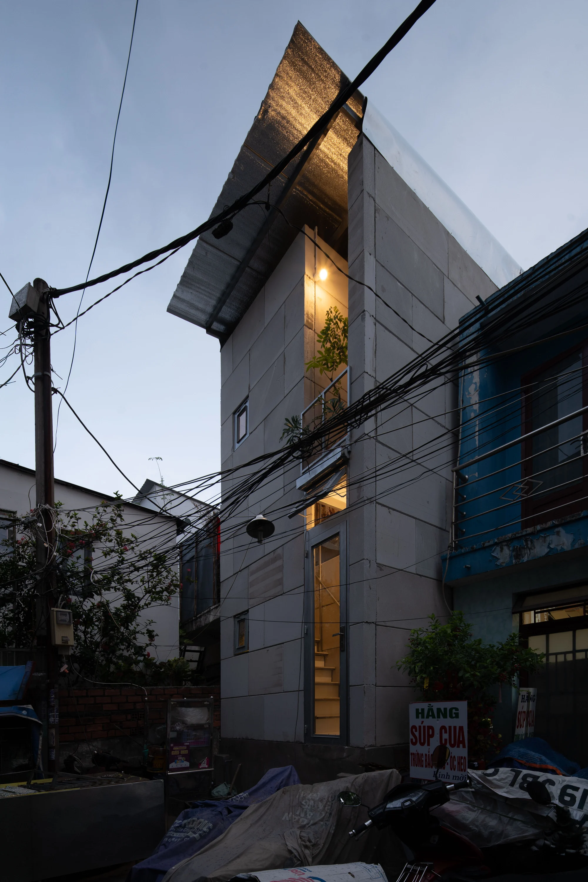 11M2 HOUSE by scale50, photographed by Nguyen Manh Toan — Dusk exterior, the house glowing warm through tangled overhead cables, the corrugated cap silhouetted against blue sky.