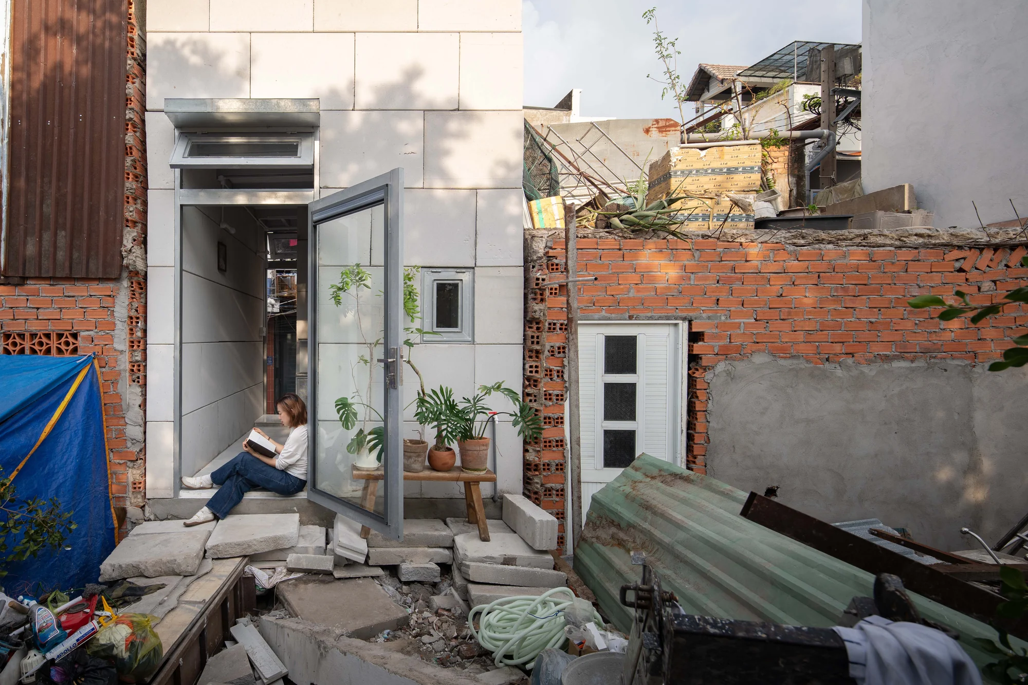 11M2 HOUSE by scale50, photographed by Nguyen Manh Toan — Ground floor entry with a young reader, glazed door open to the lane, a monstera and a low timber bench against pale AAC walls.