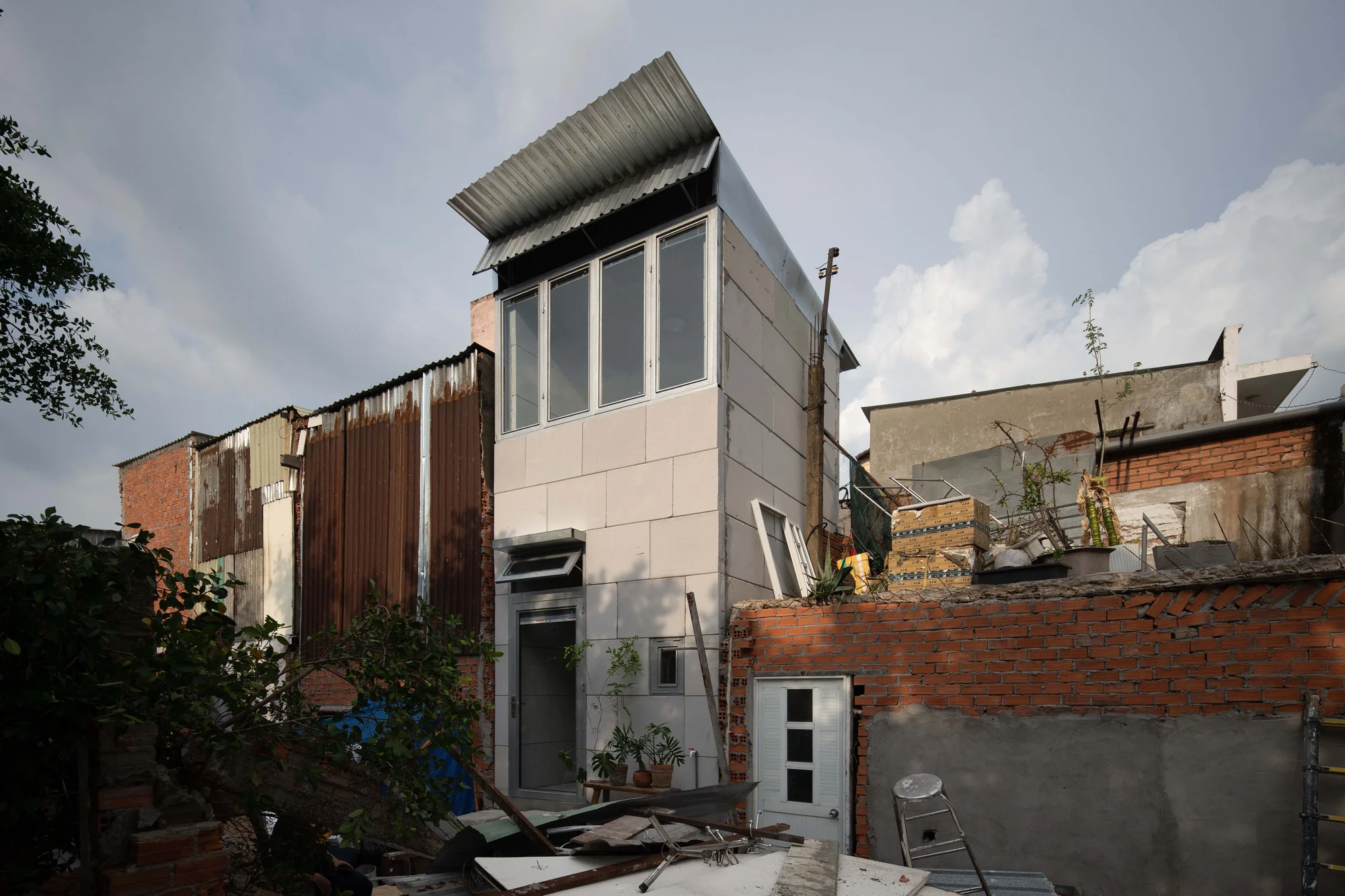 11M2 HOUSE by scale50, photographed by Nguyen Manh Toan — Facade at midday, four-bay window over a flat panel skin, set between rusted corrugated metal and raw brick of the neighbouring lots.