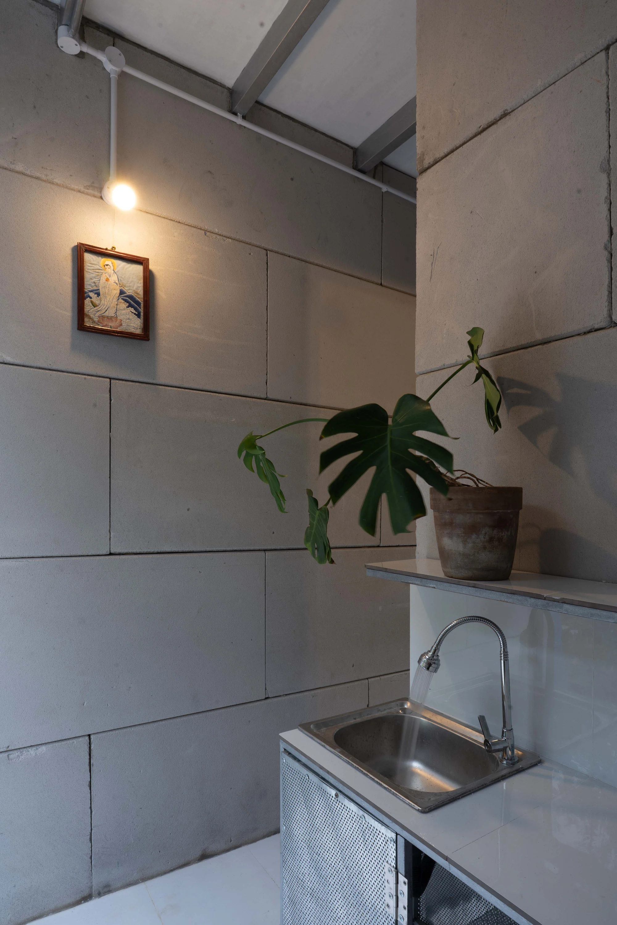 11M2 HOUSE by scale50, photographed by Nguyen Manh Toan — Kitchen corner with a galvanised steel sink, wall sconce, framed print and a monstera leaning into the light.