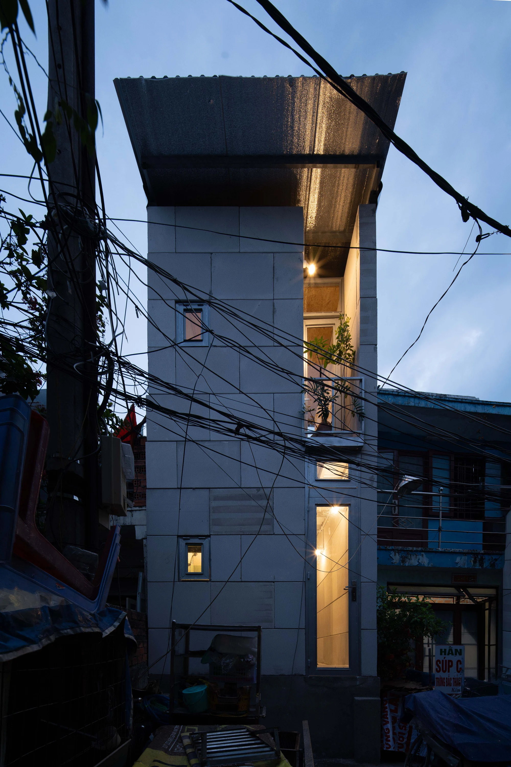 11M2 HOUSE by scale50, photographed by Nguyen Manh Toan — Blue-hour straight-on view of the slender white tower, three lit windows stacked above the entrance, neighbouring shopfronts dark to either side.