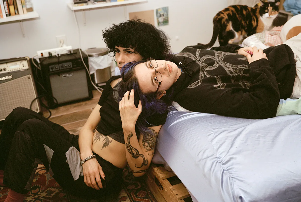 Christopher Postlewaite by Christopher Postlewaite, photographed by Christopher Postlewaite — Two people lying tangled on a low mattress against a guitar amp and Persian rug, a calico cat watching from the rumpled bedding behind them.