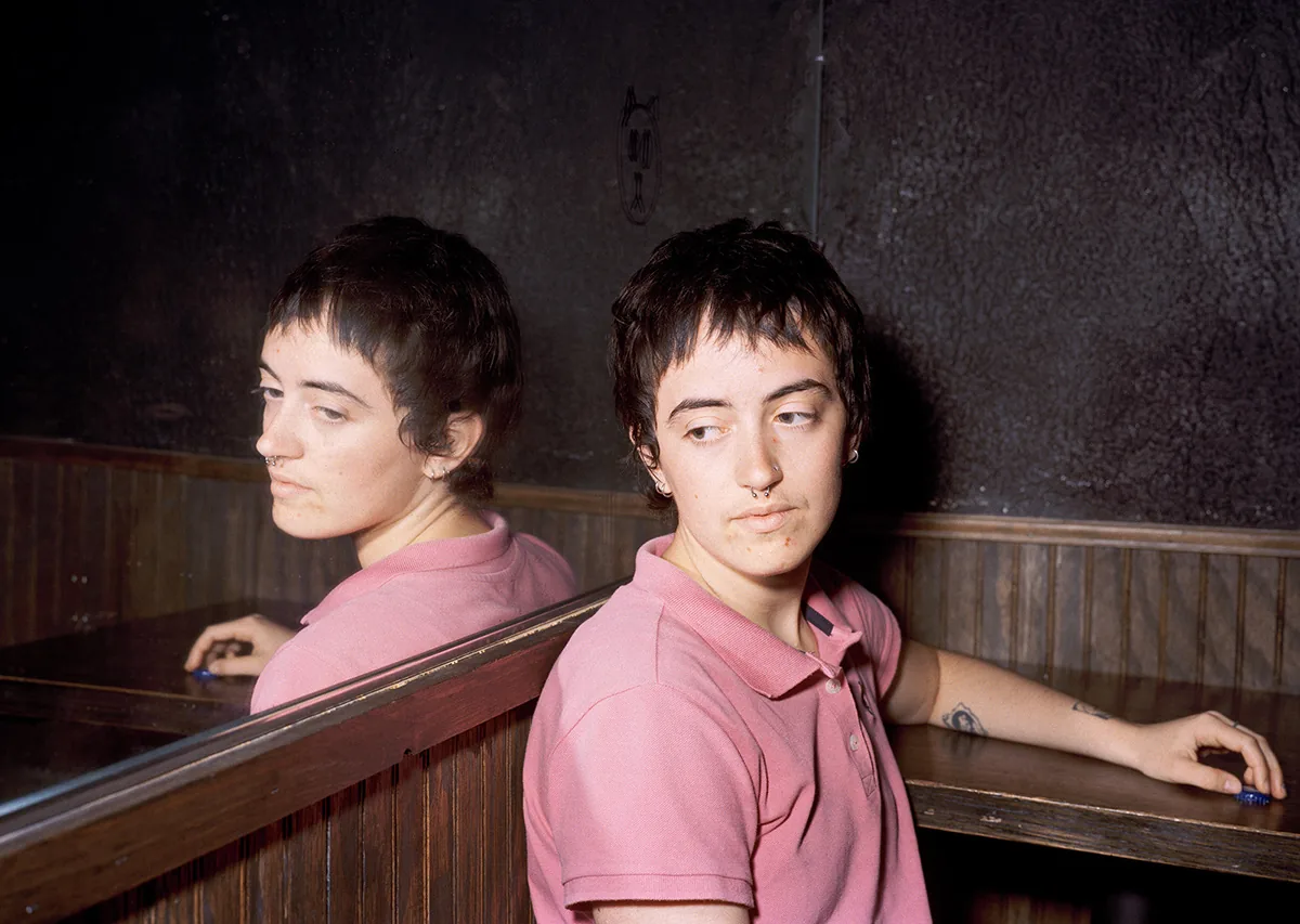 Christopher Postlewaite by Christopher Postlewaite, photographed by Christopher Postlewaite — A young woman in a pink polo shirt seated next to her mirror reflection inside a wood-panelled bar booth, twin nose rings catching the low overhead light…