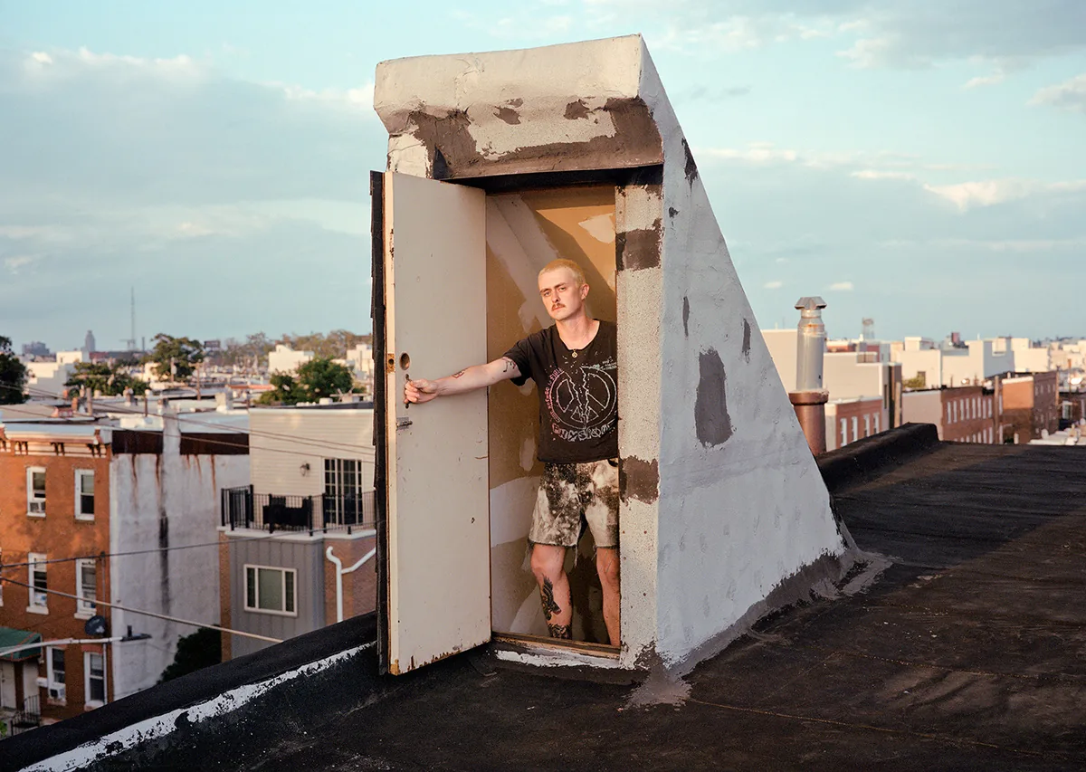 Christopher Postlewaite by Christopher Postlewaite, photographed by Christopher Postlewaite — A figure standing inside a rooftop bulkhead door, the open white door swung wide and Philadelphia's row-house skyline stretching to the horizon at golden hour.