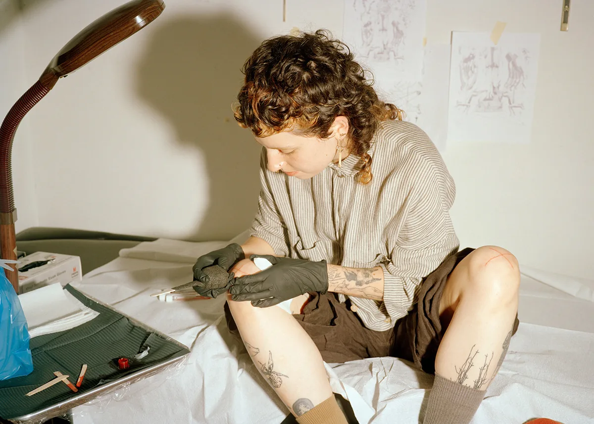 Christopher Postlewaite by Christopher Postlewaite, photographed by Christopher Postlewaite — A tattoo artist in black gloves working on a sitter's outer thigh, plant and figure studies pinned to the wall behind, a magnifying lamp tilted in from the left.