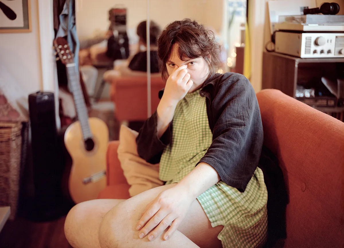 Christopher Postlewaite by Christopher Postlewaite, photographed by Christopher Postlewaite — A subject in a green-checked dress and dark linen shirt pinching the bridge of the nose on a red couch, a classical guitar and stacked hi-fi gear behind in the…