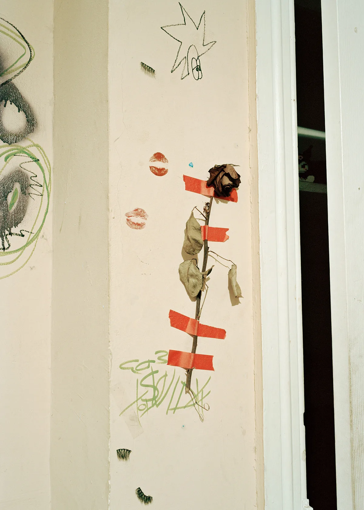 Christopher Postlewaite by Christopher Postlewaite, photographed by Christopher Postlewaite — A dried single-stem rose taped vertically to a cream interior wall with red gaffer tape, kiss-prints in two shades of pink and a green marker scrawl below.