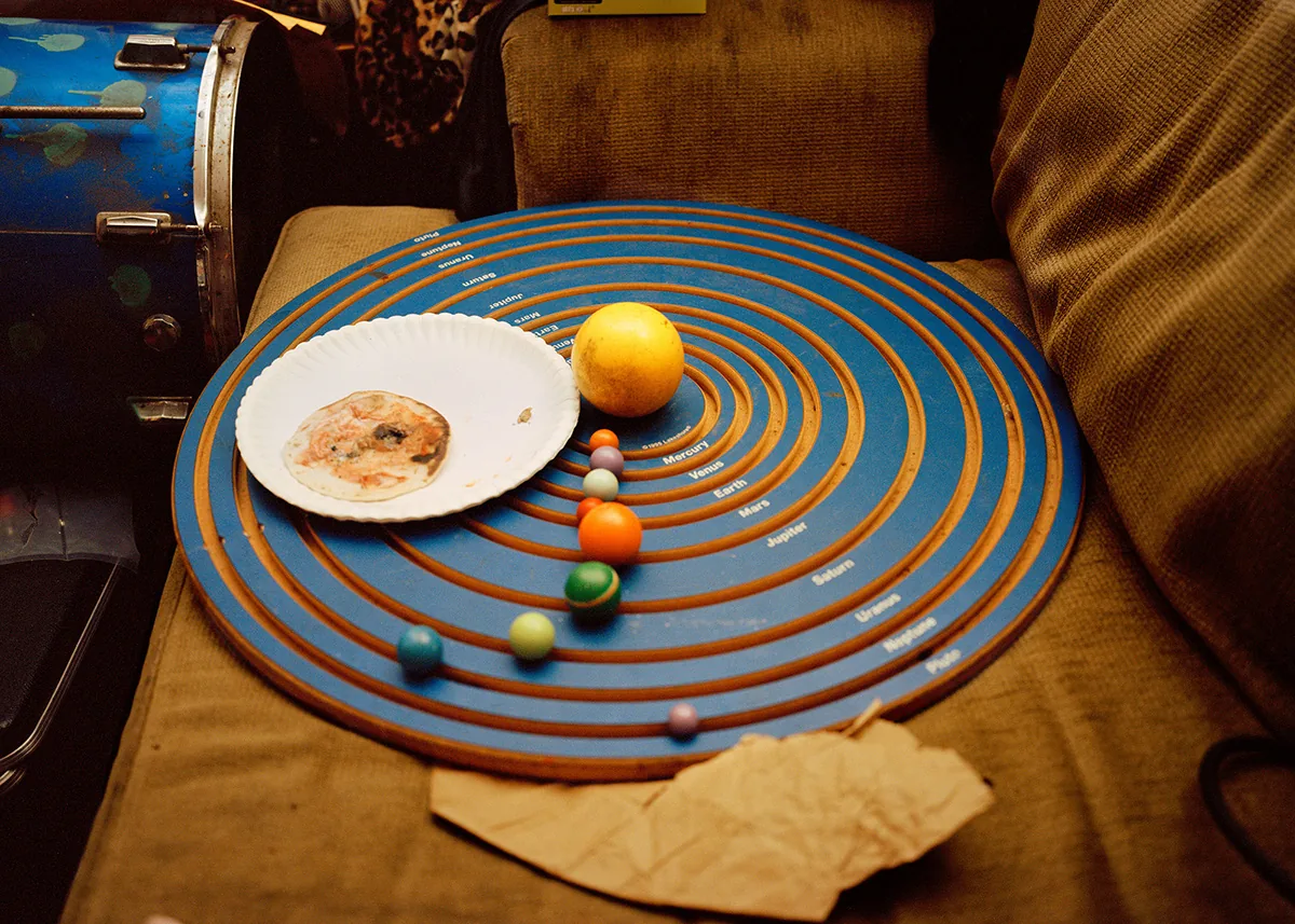 Christopher Postlewaite by Christopher Postlewaite, photographed by Christopher Postlewaite — A blue cardboard solar-system game board placed on a brown velour couch with a half-eaten donut on a paper plate at Mercury and coloured marbles tracking the…