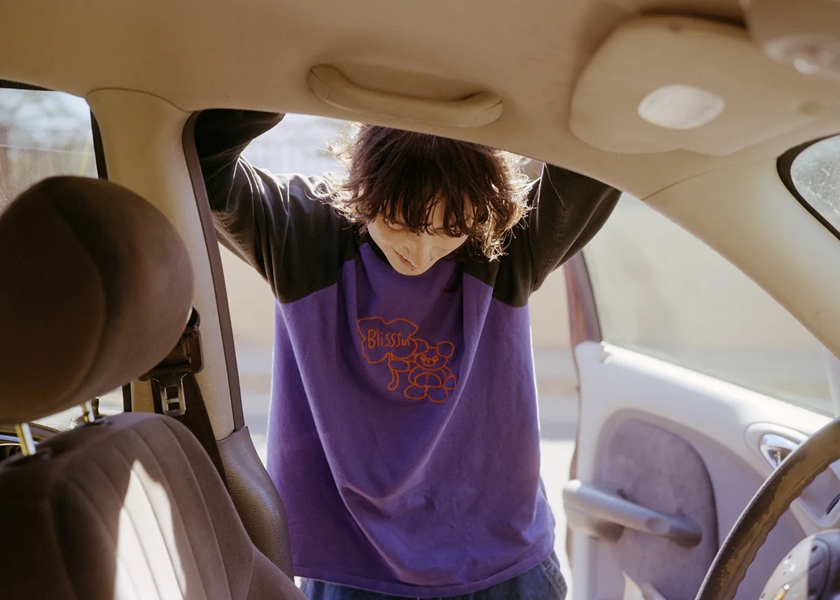 Christopher Postlewaite by Christopher Postlewaite, photographed by Christopher Postlewaite — A figure leaning into the open door of a sun-warmed sedan, a purple Blissful t-shirt with an orange teddy-bear graphic catching the dust-light of the cabin.