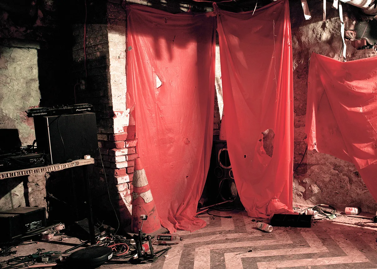 Christopher Postlewaite by Christopher Postlewaite, photographed by Christopher Postlewaite — Interior of the after-hours bar Foto Club before the April fire: red sheer curtains draped over a Pioneer DJ rig, traffic cone, exposed brick and a checkered…