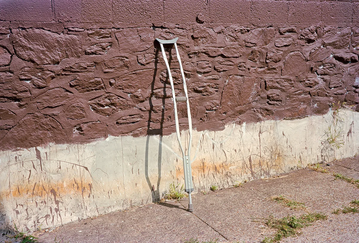 Christopher Postlewaite by Christopher Postlewaite, photographed by Christopher Postlewaite — A pair of aluminium crutches leaning against a stone-textured masonry wall painted half-burgundy, half-cream, midday shadow falling across cracked pavement at…
