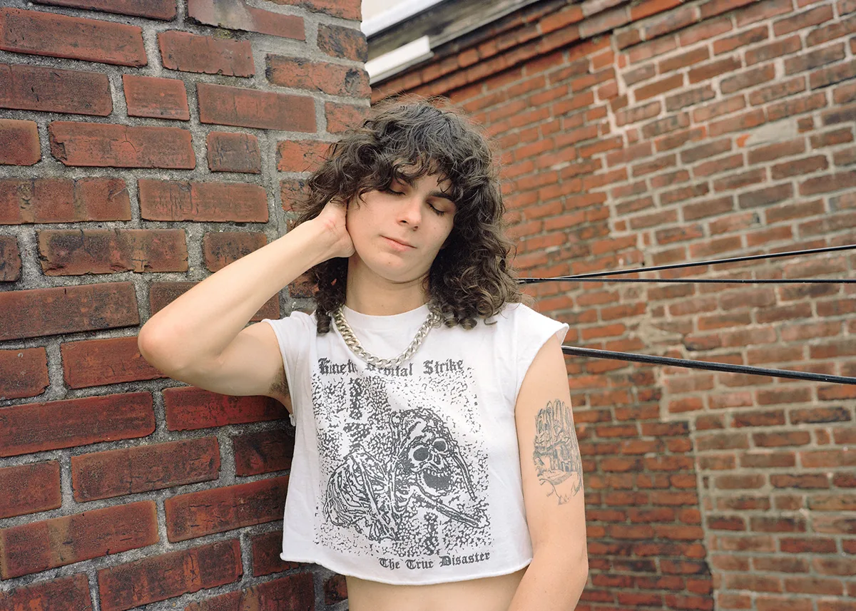 Christopher Postlewaite by Christopher Postlewaite, photographed by Christopher Postlewaite — A young person in a Mental Strike skull-graphic crop top leaning against a sunlit red brick wall, eyes closed, silver chain at the neck, snake tattoo across…