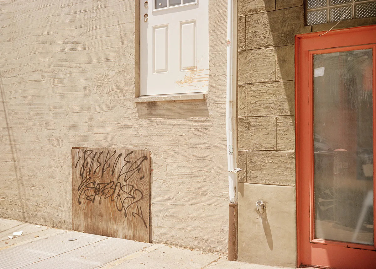 Christopher Postlewaite by Christopher Postlewaite, photographed by Christopher Postlewaite — A graffiti-tagged plywood square fitted under a white-painted door, a red shopfront and a downpipe sharing the same patch of bleached stucco sidewall.