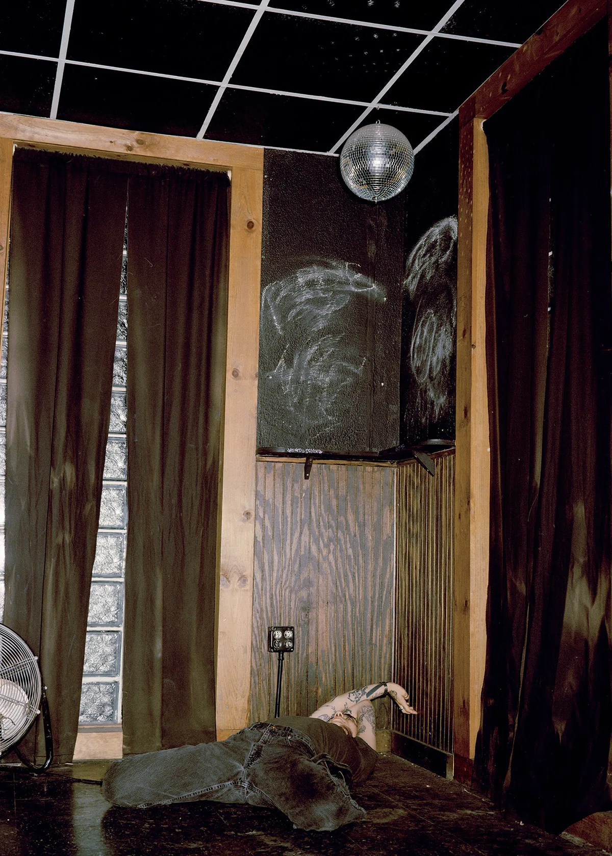 Christopher Postlewaite by Christopher Postlewaite, photographed by Christopher Postlewaite — A figure laid out on the floor of a curtained back room with a small disco ball overhead and chalk-drawn faces above the wood-panelled wainscot.