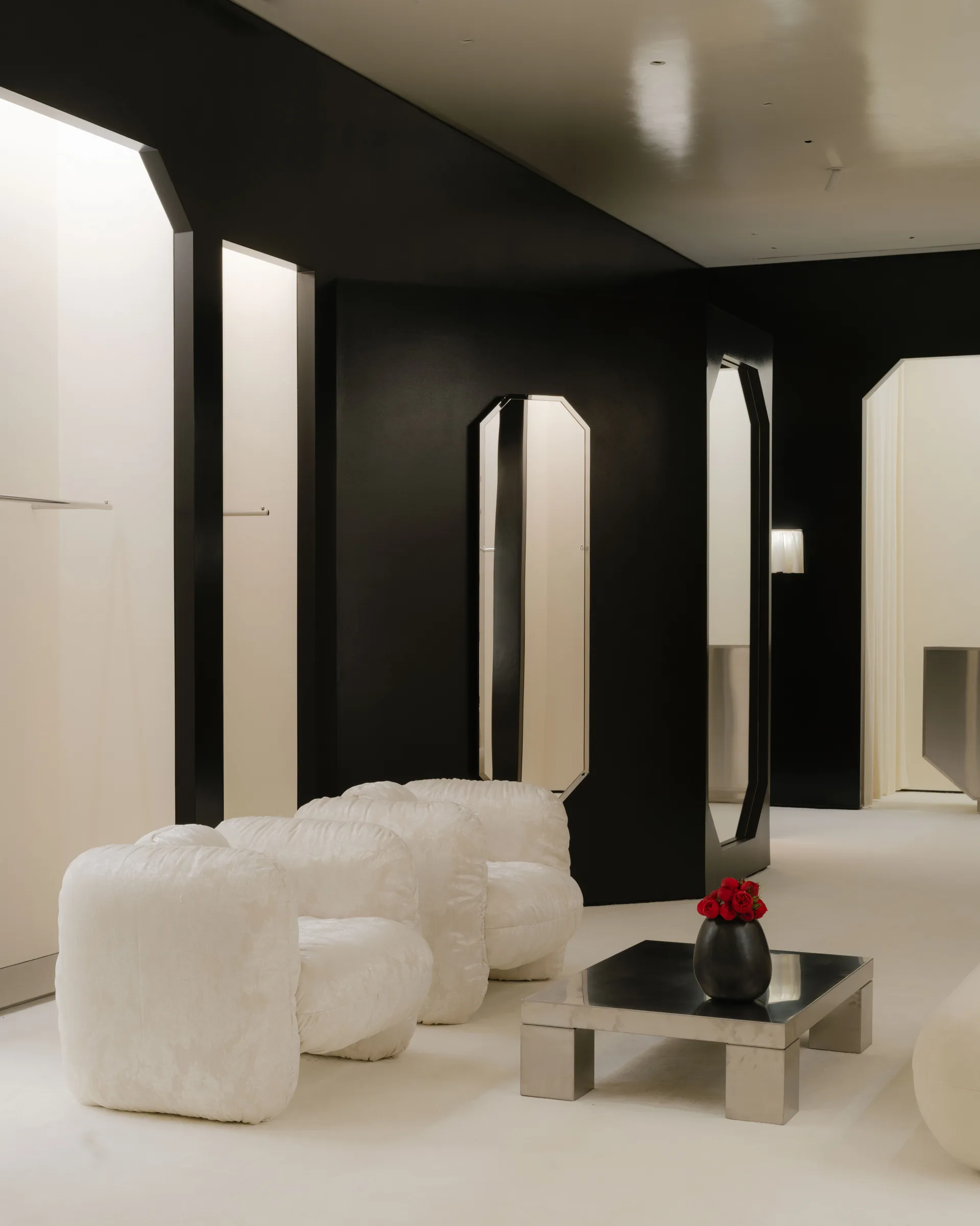 Roses Lace by EDXXKAT — Three-quarter view of the lounge corner with three octagonal black mirrors leaned against the black panel, two white shearling pouf-cubes in the foreground and a brushed-metal coffee table with a single red rose at centre.