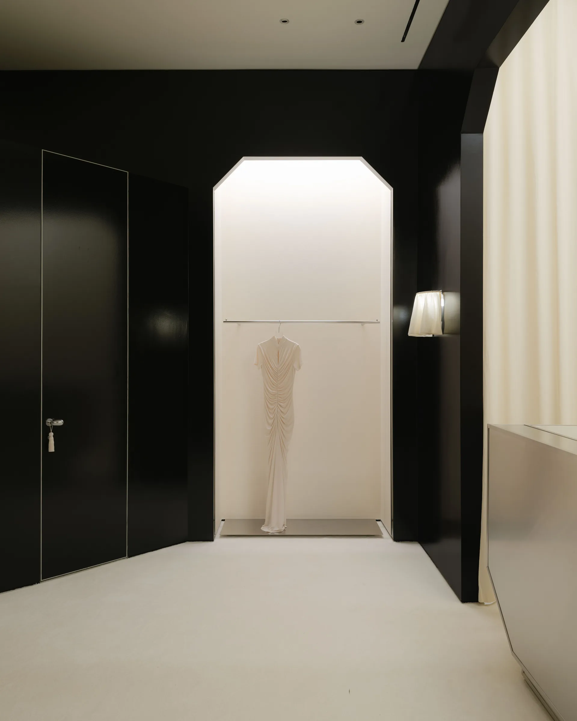Roses Lace by EDXXKAT — Tall lightbox niche framing a draped ivory column dress on a brass rail, the glossy black surround reflecting the niche's light back onto the floor, a concealed black door visible to the left.