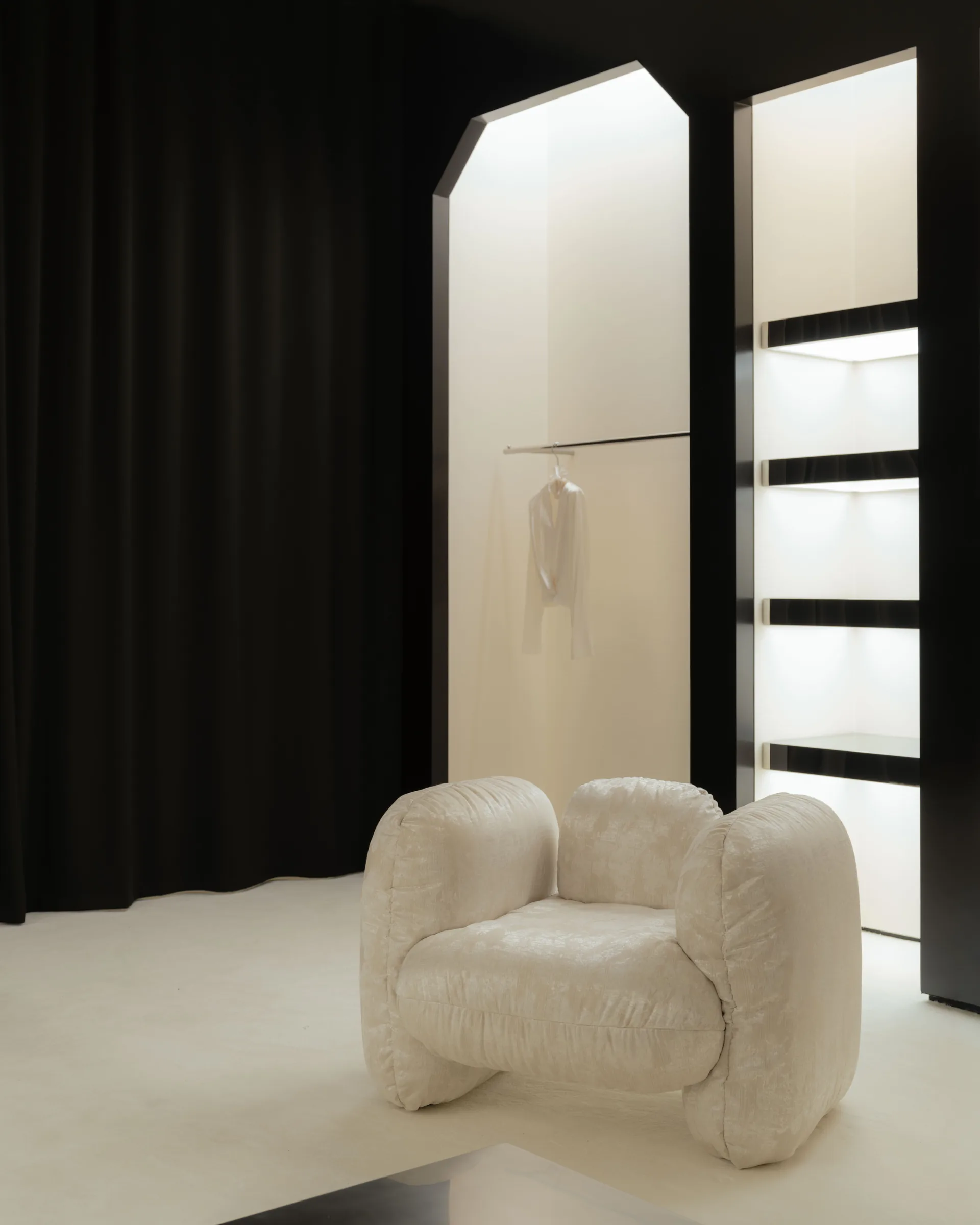 Roses Lace by EDXXKAT — Detail of a single cream textured armchair with rounded volumes, set in front of a tall lightbox niche holding a hanging dress; a slim glass shelving column glows to the right.
