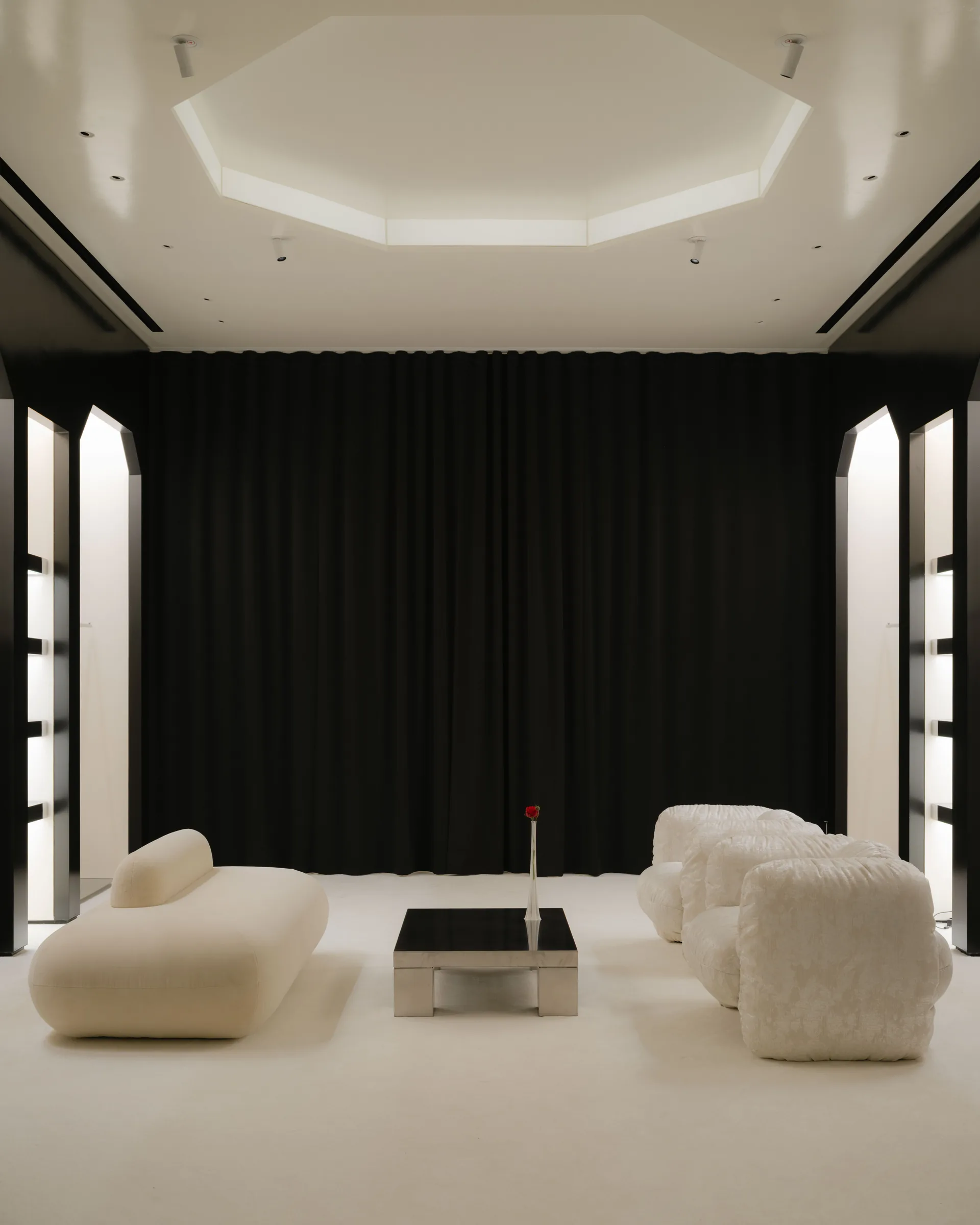 Roses Lace by EDXXKAT — Centred view of the lounge with the black stage curtain pulled across the back wall: white shearling poufs face a curved cream sofa across a brushed-metal coffee table holding a single rose, octagonal coffer above lit in white.