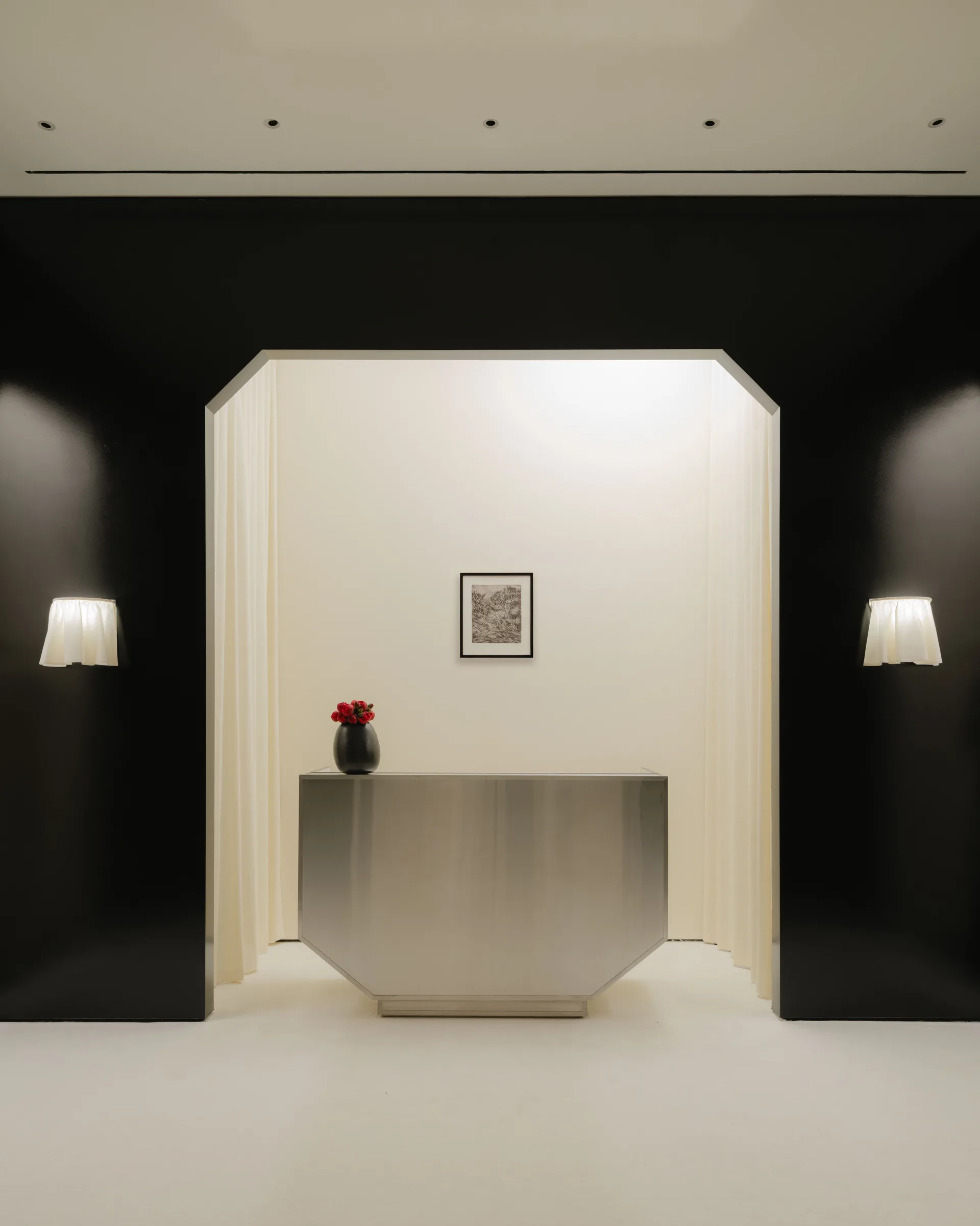 Roses Lace by EDXXKAT — Symmetrical elevation of the cash desk: the brushed stainless steel volume sits inside a tall white-curtained recess, twin pleated sconces flanking it on the surrounding black walls.