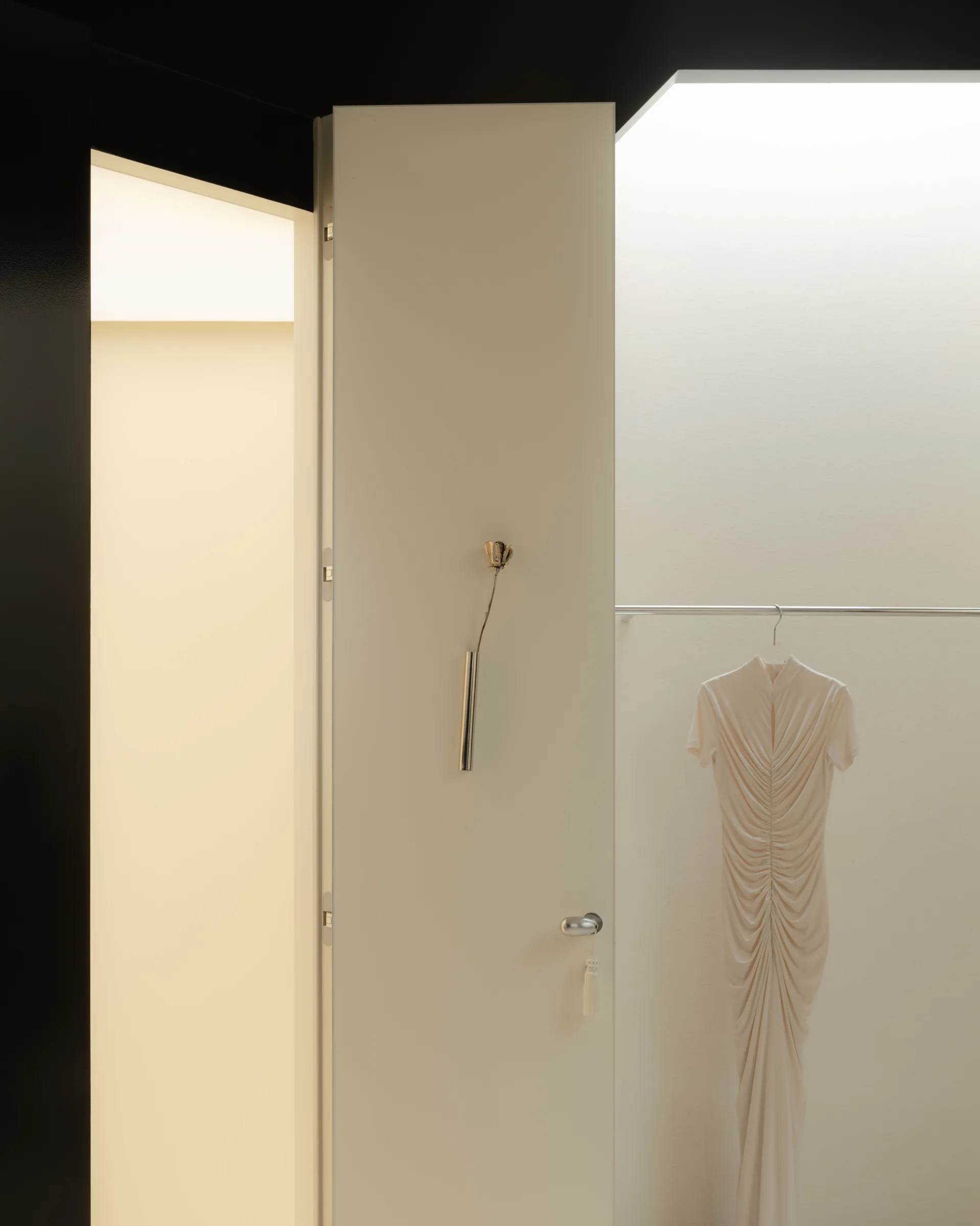 Roses Lace by EDXXKAT — Detail of a cream-painted fitting room threshold: a brushed-brass shoehorn and door catch set into a recessed panel, an ivory dress visible hanging in the lit niche beyond.