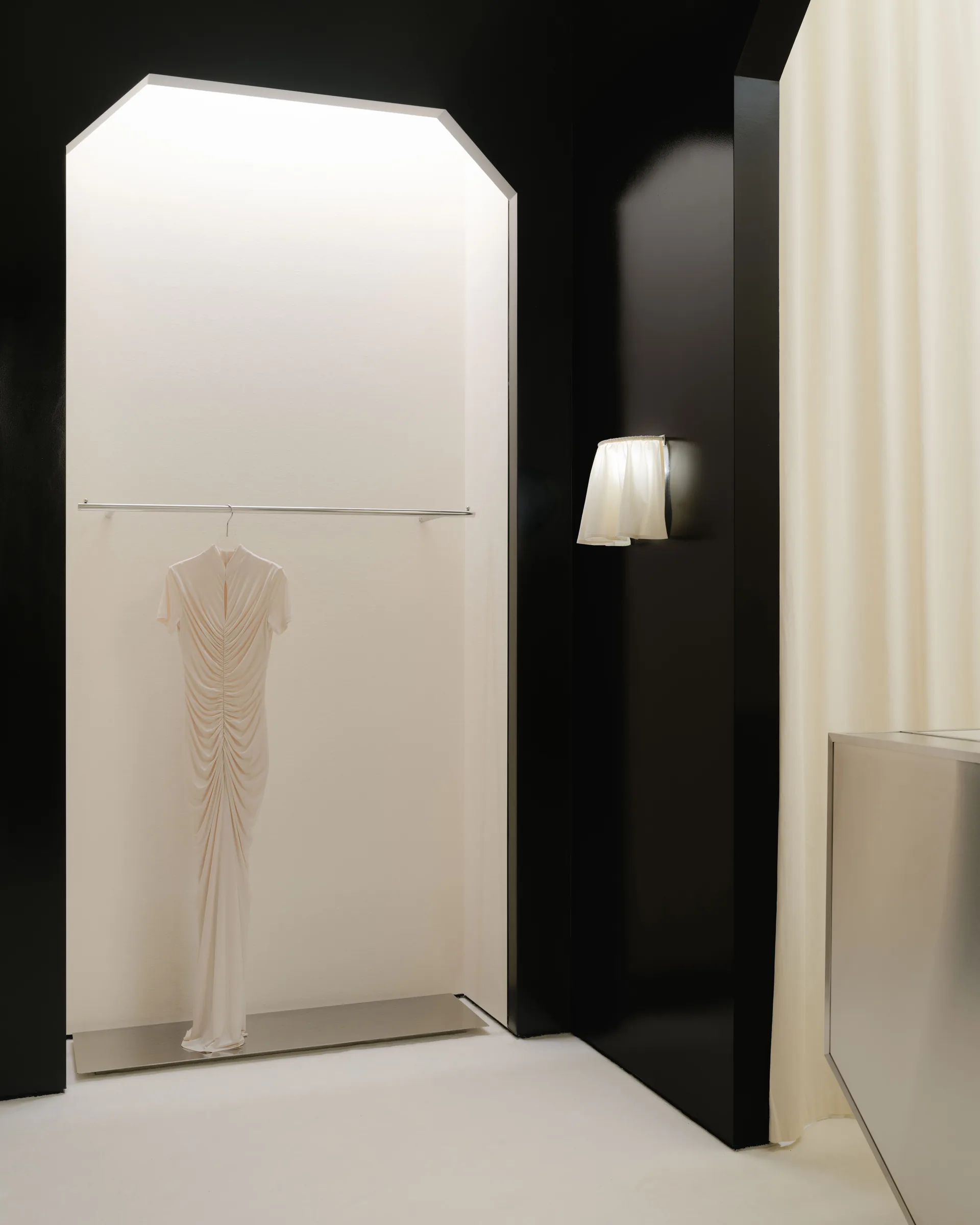 Roses Lace by EDXXKAT — Close-up of a single ruched ivory dress hanging from a brass rail inside an illuminated white niche, set into the surrounding glossy black wall, a pleated cream sconce glowing to the right.