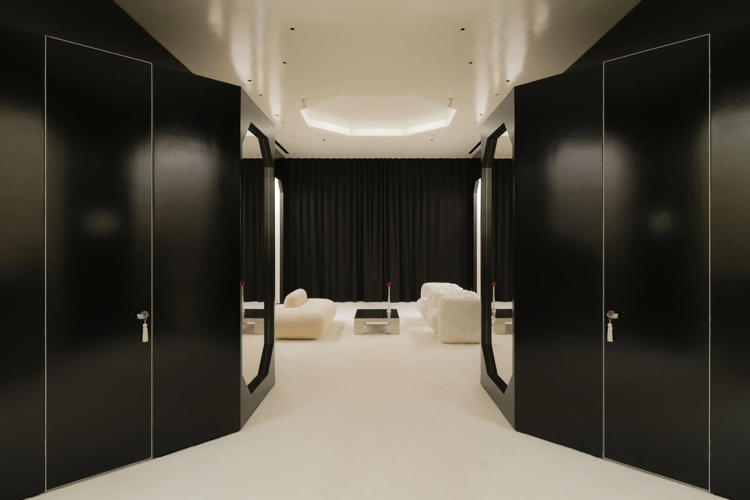 Roses Lace by EDXXKAT — View from a corridor between two glossy black sliding panels into the lounge: shearling poufs and a low table arrayed against the black stage curtain, the octagonal lit coffer overhead just visible at the top of the frame.