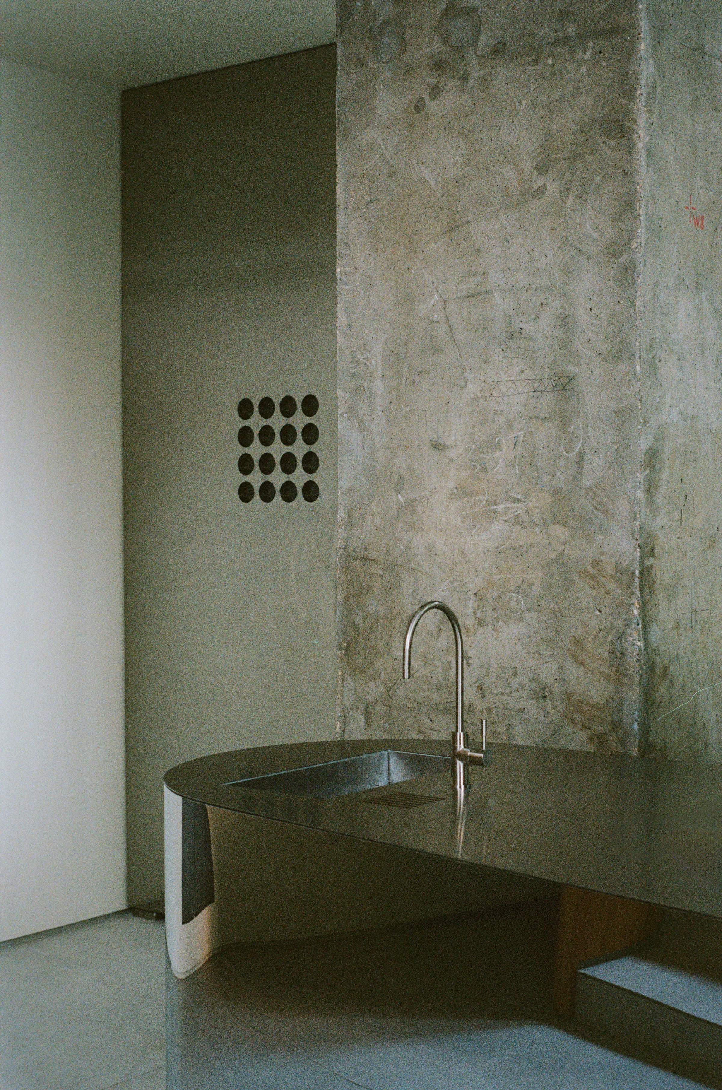 Nothing Fancy by EDXXKAT — Detail at the steel-topped counter: a stainless steel tap rises from a brushed steel surface set against a raw concrete wall punched with a small grid of dark dots, the floor in polished cement.