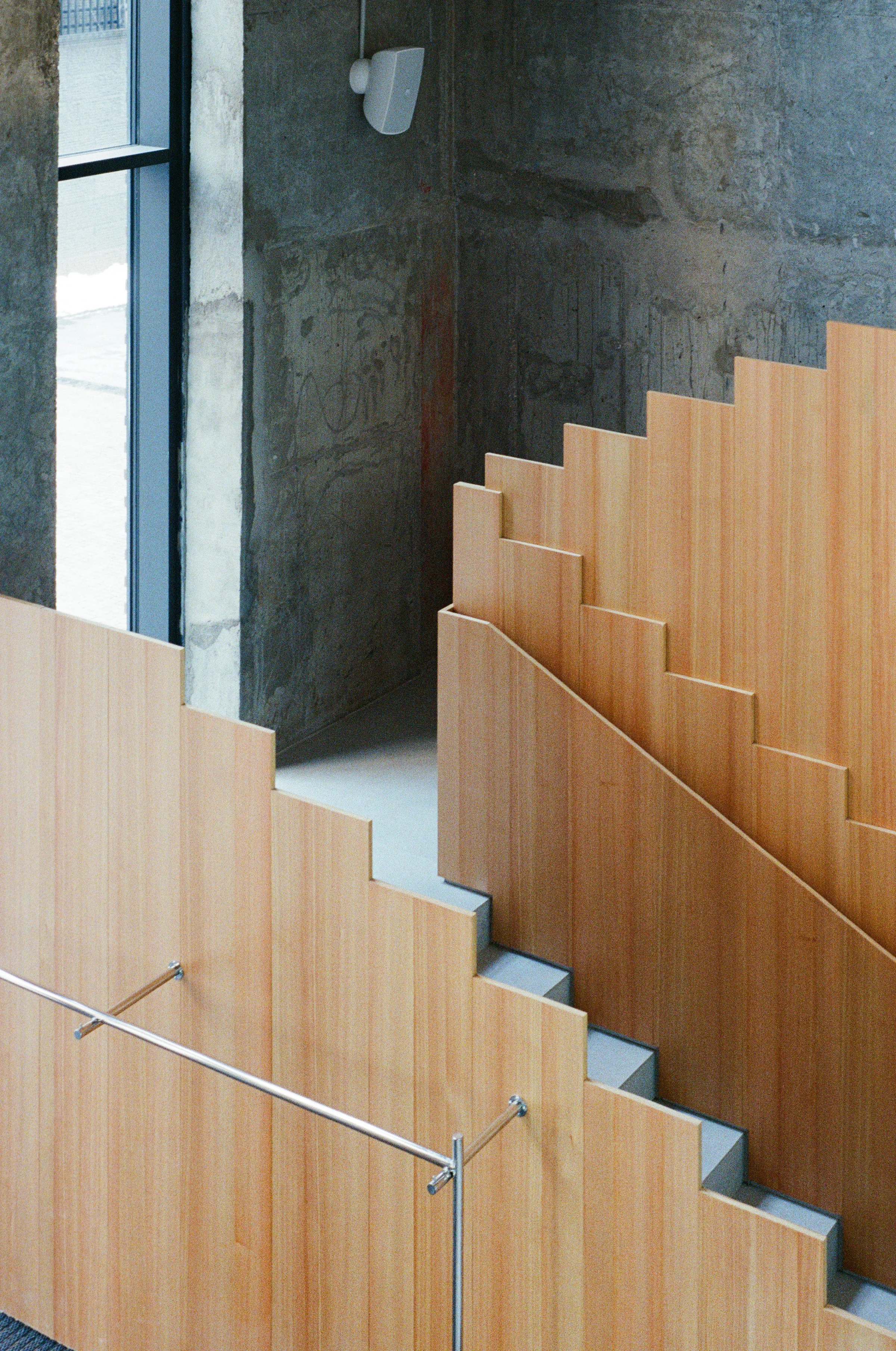 Nothing Fancy by EDXXKAT — Larch staircase folded against a raw concrete wall: stepped planar treads in pale wood, a slim stainless steel handrail bracketed off the wood, a tall window casting daylight onto the patinated concrete behind.