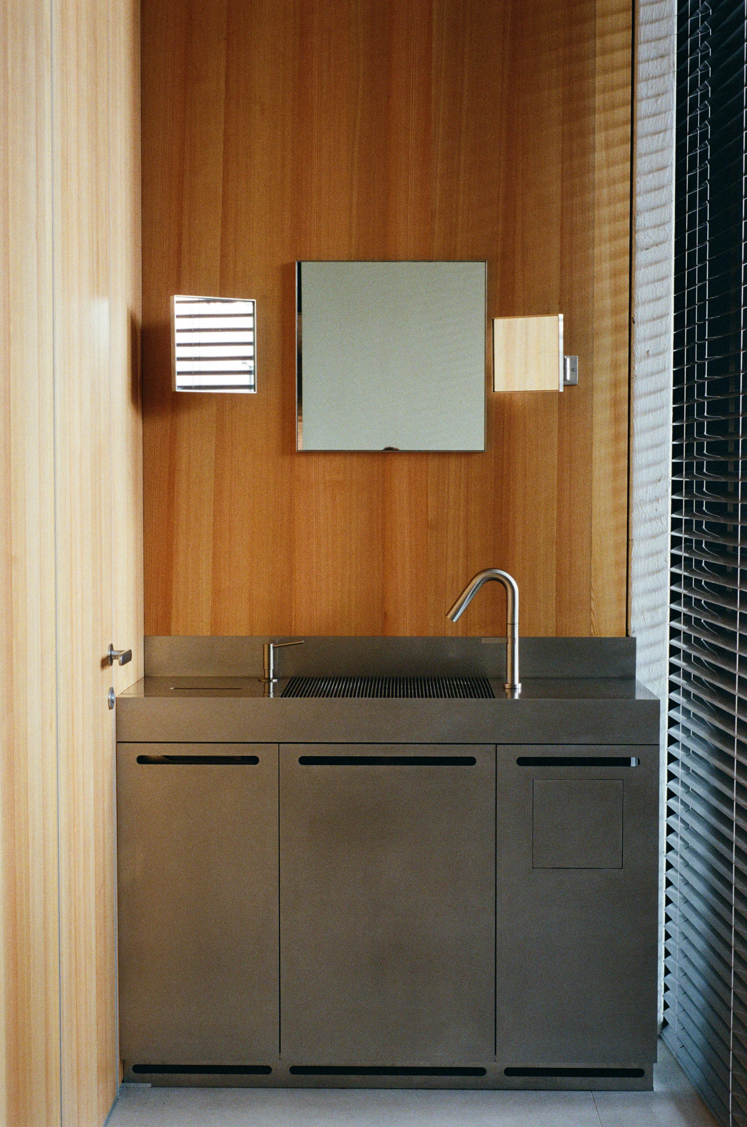 Nothing Fancy by EDXXKAT — Washroom corner: stamped stainless steel sink unit set into a vertical larch panelled wall, a small square mirror flanked by two wall sconces, a venetian blind to the right catching daylight.