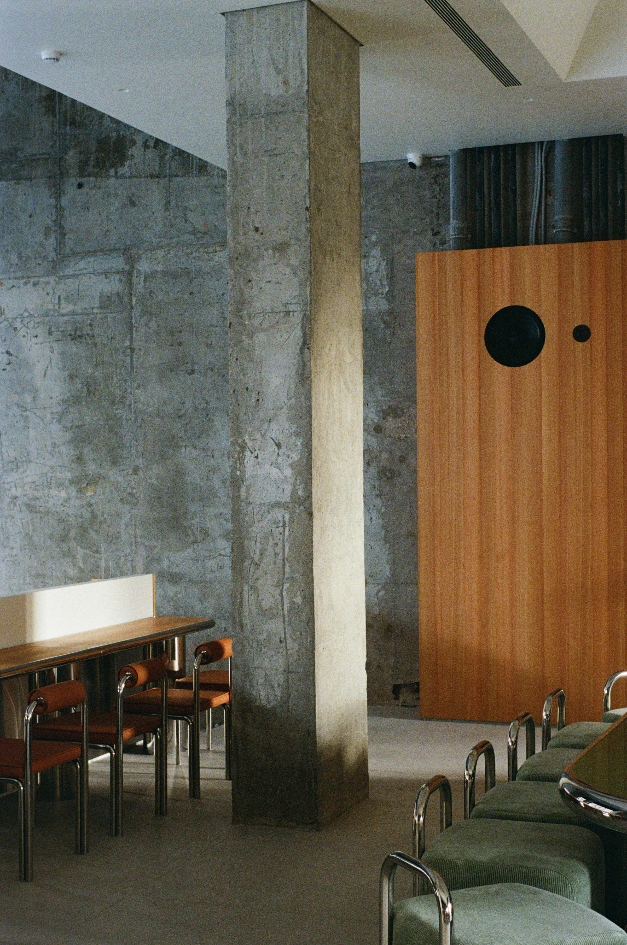 Nothing Fancy by EDXXKAT — Wide cafe view: a raw concrete column with board-marks and patina holds the centre, brown leather chairs on tubular steel frames at the left, a deep green velvet sofa at the right, a larch veneer wall with a black circular…