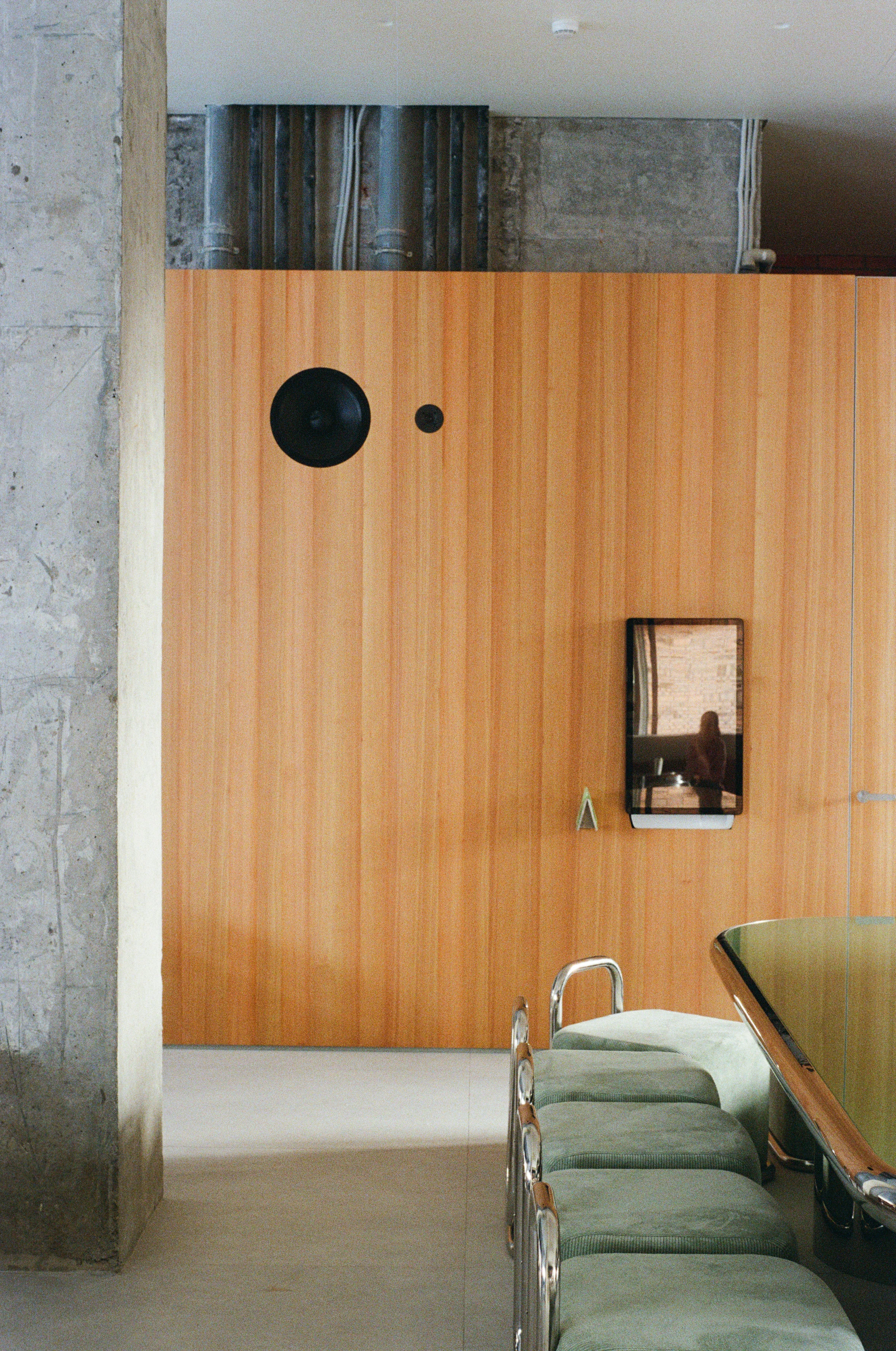 Nothing Fancy by EDXXKAT — Material contact: a vertical larch veneer wall with a large and small black porthole punched through it, the building's exposed ductwork running above, a row of green velvet bench backs visible at the bottom right.