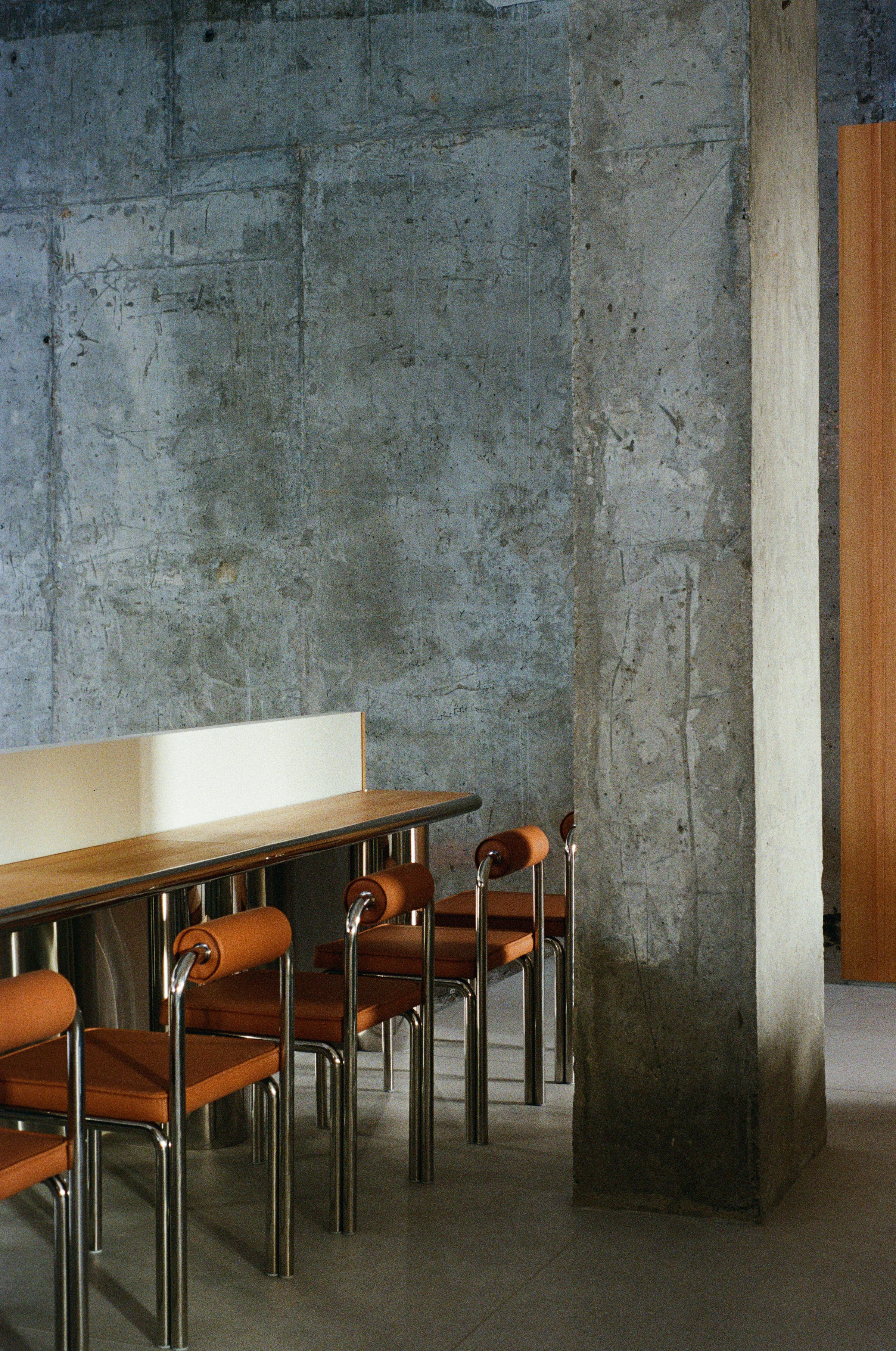Nothing Fancy by EDXXKAT — Concrete column close-up: pour seams and chalked construction marks left fully visible, a row of brown leather chairs on tubular steel legs at left, a deep green velvet armchair frame at right.