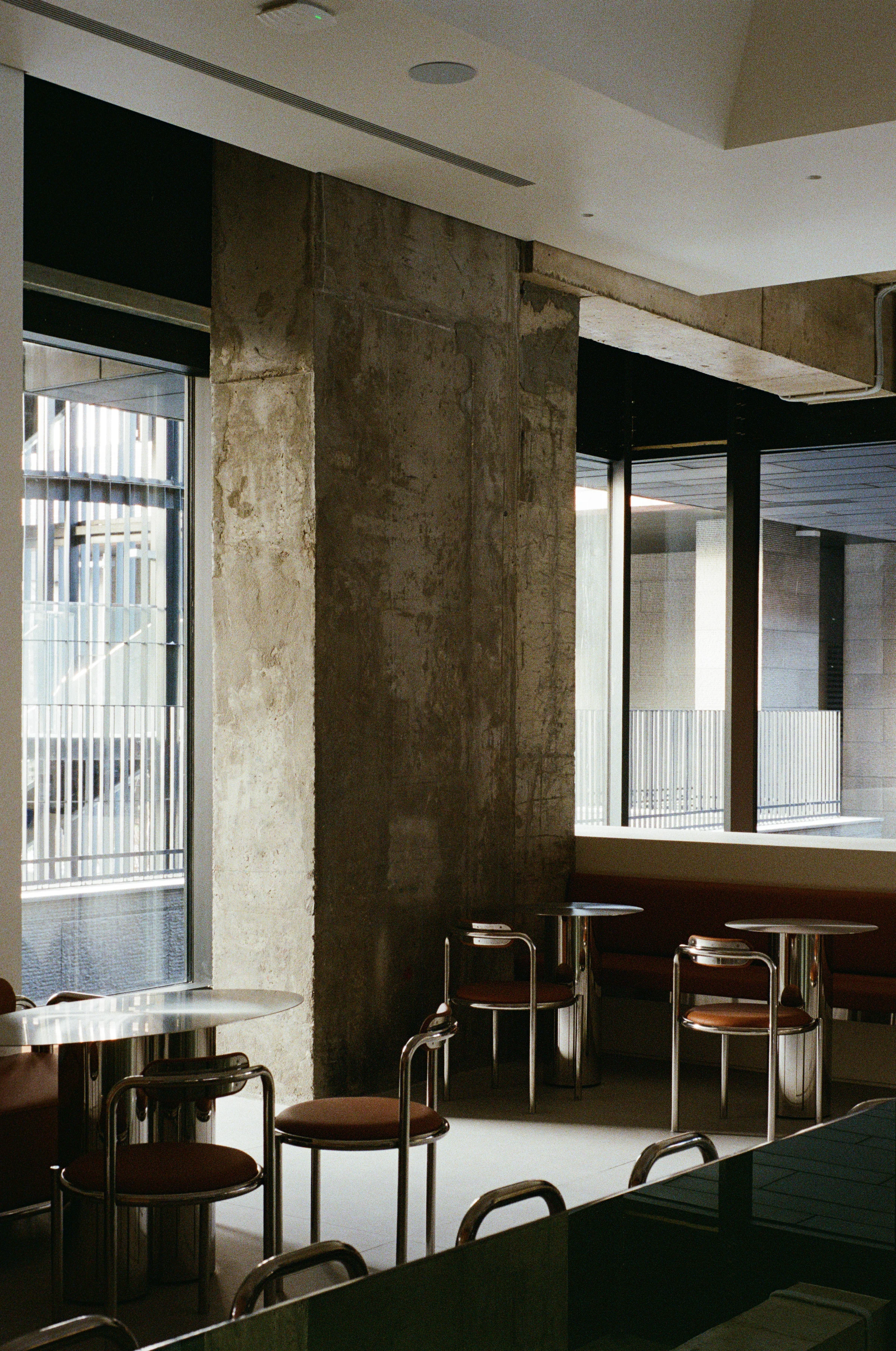 Nothing Fancy by EDXXKAT — Window bay: brown leather banquette running below a wide black-framed window, polished stainless steel side tables with marble tops, tubular steel chairs with leather seats, a raw concrete soffit overhead.