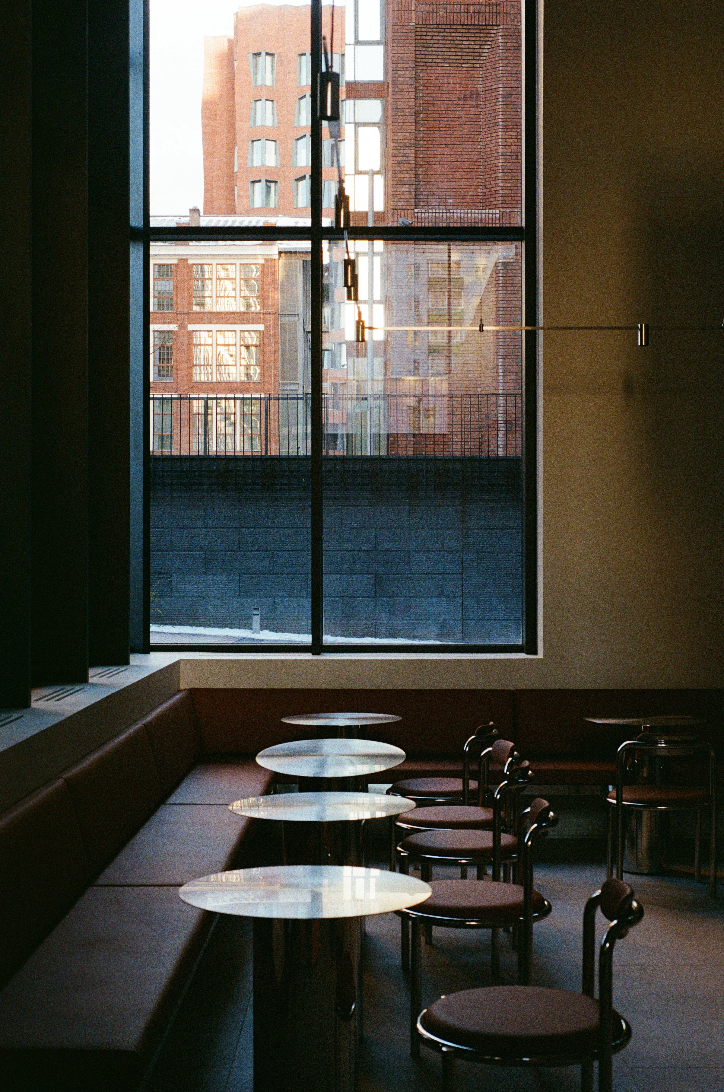 Nothing Fancy by EDXXKAT — View across a steel-edged marble counter to a wall of glazing, the neighbouring red brick building visible outside, pendant lights on a horizontal track, leather banquette wrapping the bay.