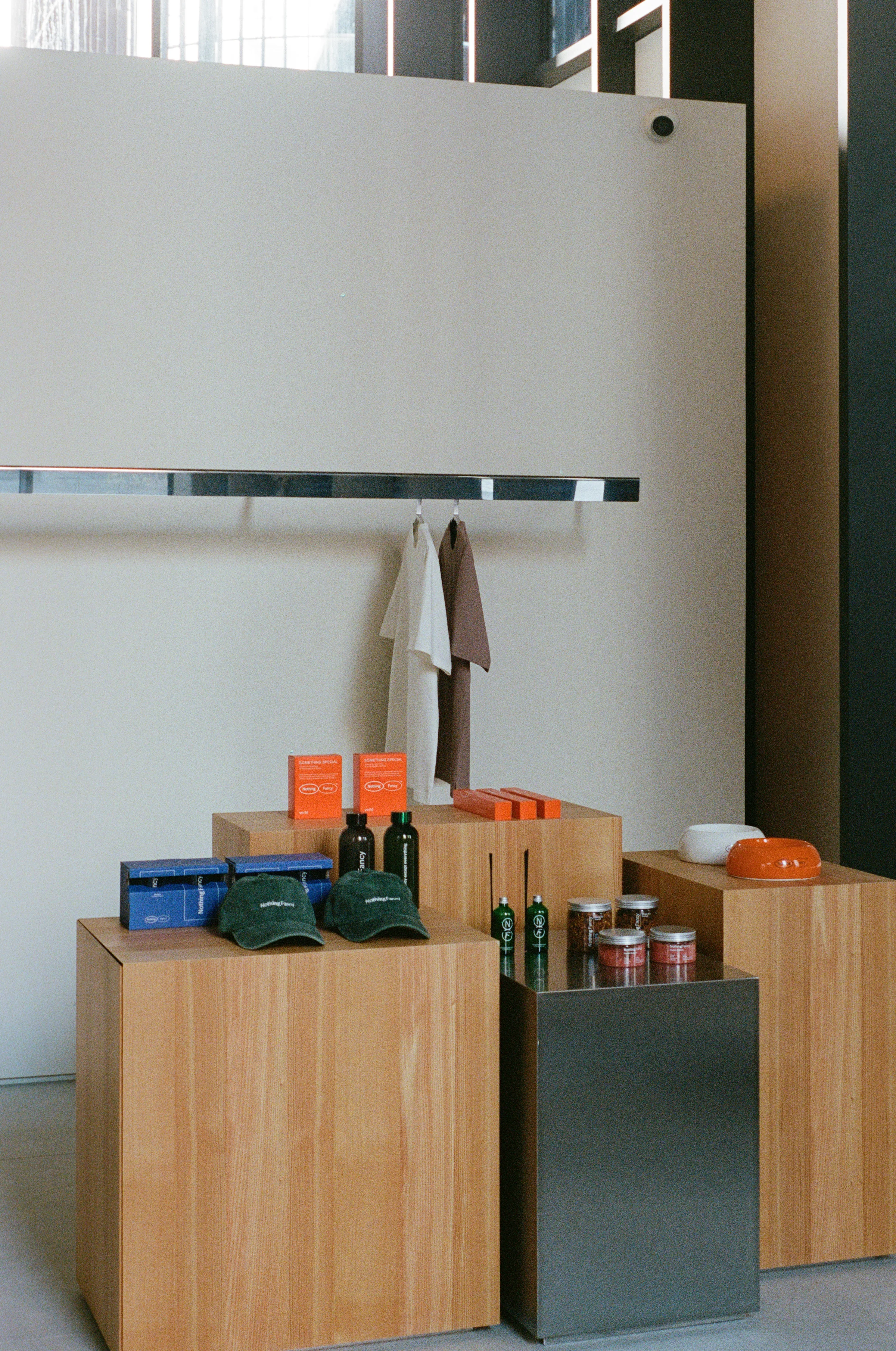 Nothing Fancy by EDXXKAT — Retail plinth: a cluster of raw larch and steel cubes display branded caps, coffee bottles and jars, a polished stainless steel rod hung with workwear above, an orange paper poster propped on a plinth at the back.
