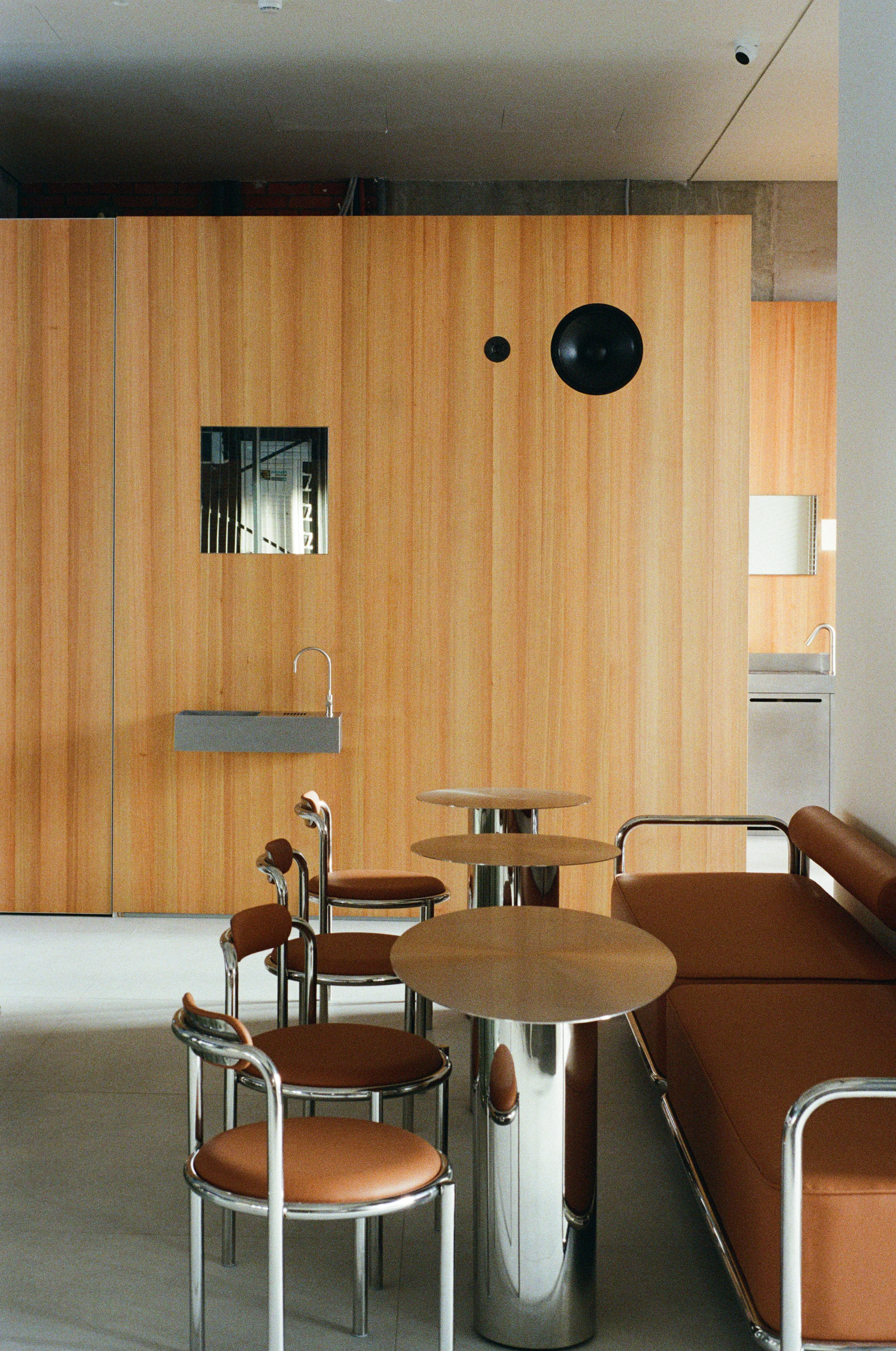 Nothing Fancy by EDXXKAT — Main room: a full-height larch veneer wall with two black portholes and an inset stainless steel basin, a long brown leather sofa to the right, marble-topped polished steel cafe tables in the foreground, raw concrete column to…