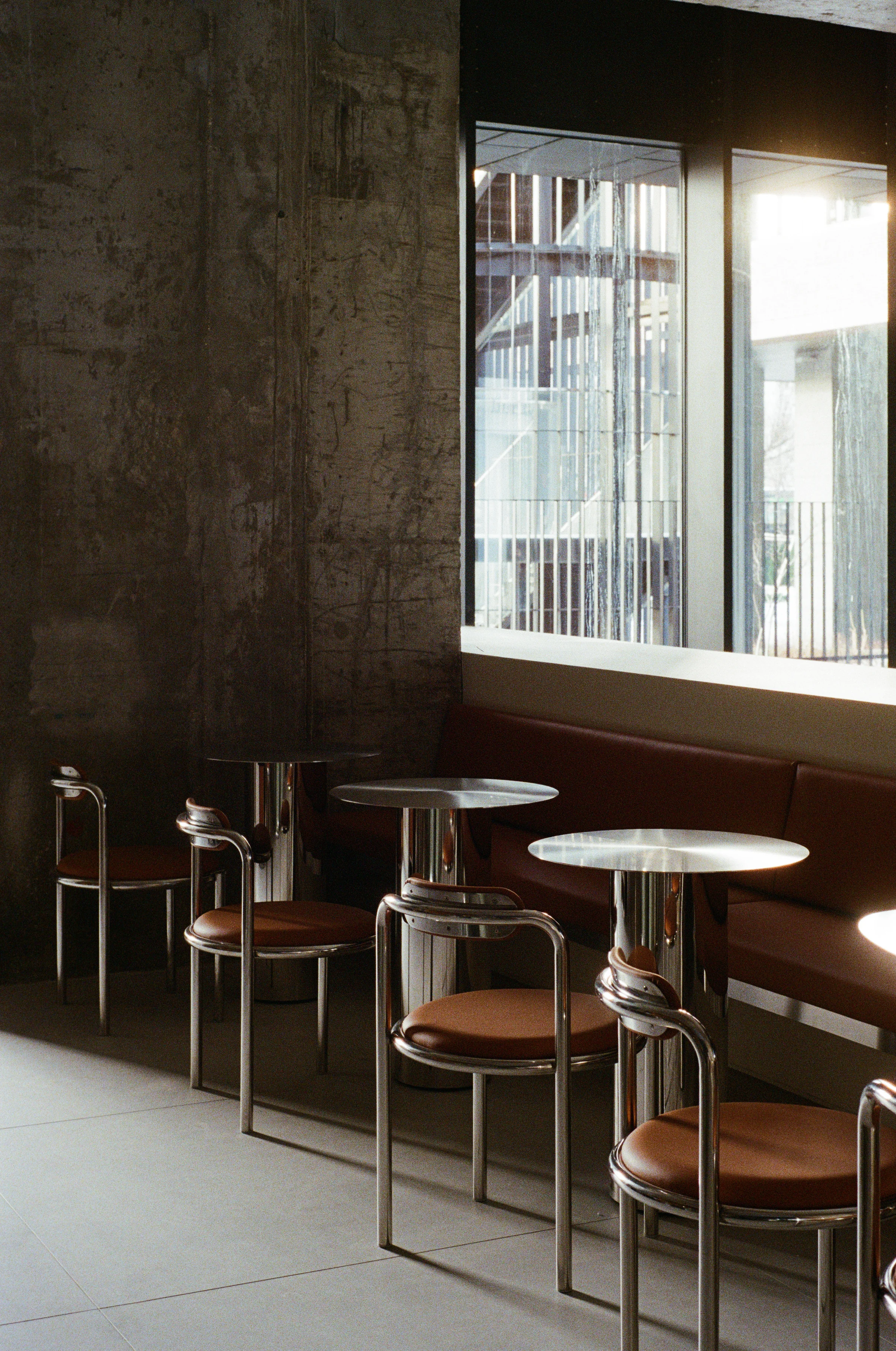 Nothing Fancy by EDXXKAT — Service nook: a larch panelled wall holds a small square mirror, a stainless steel hand-basin, and a black circular porthole; a green glass communal table edge runs in the foreground with venetian blinds at the right.