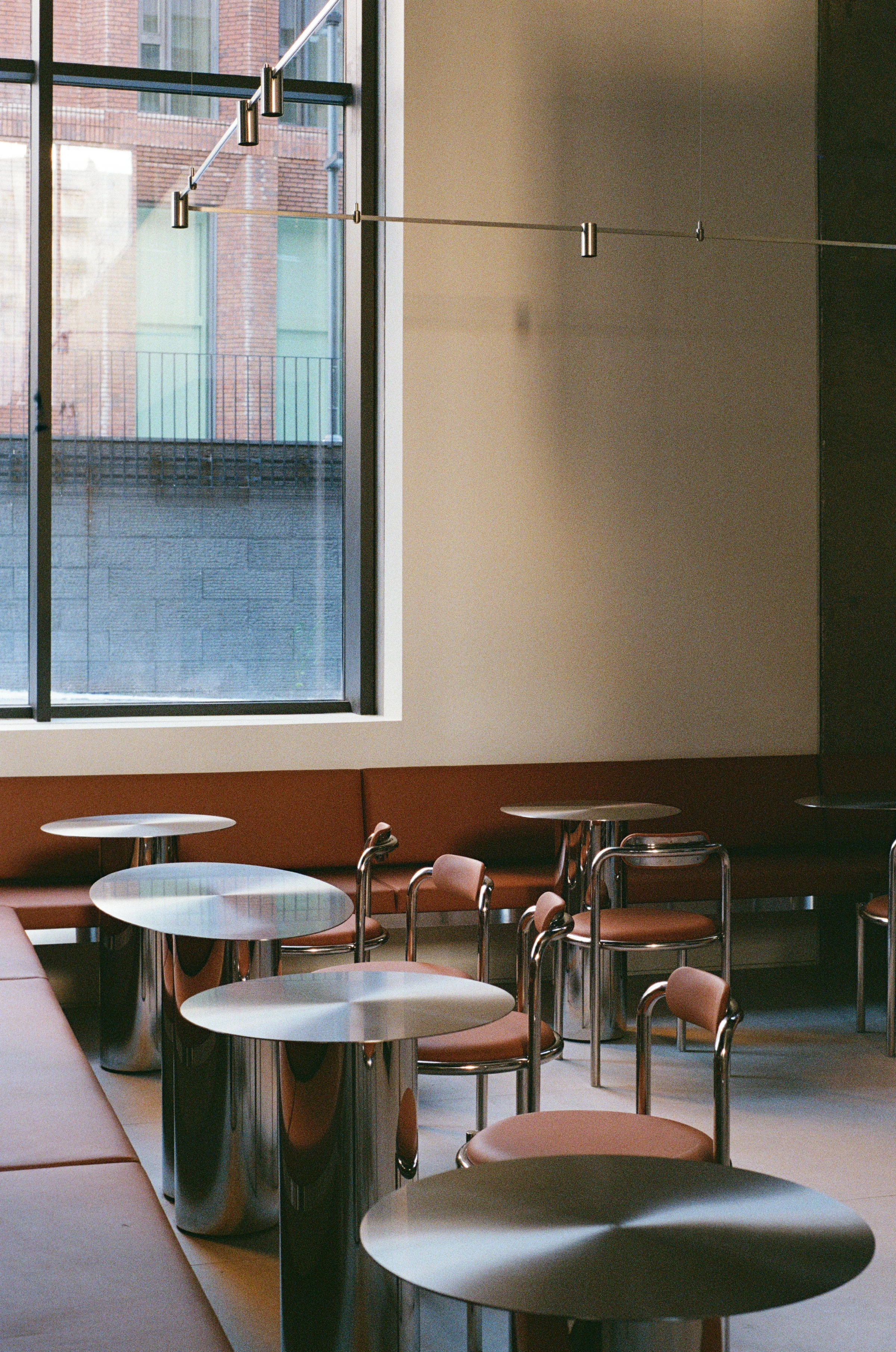 Nothing Fancy by EDXXKAT — Cafe seating row: a long brown leather banquette runs along the wall, polished stainless steel side tables with marble disc tops, tubular steel chairs with leather seats, late daylight raking across the floor.