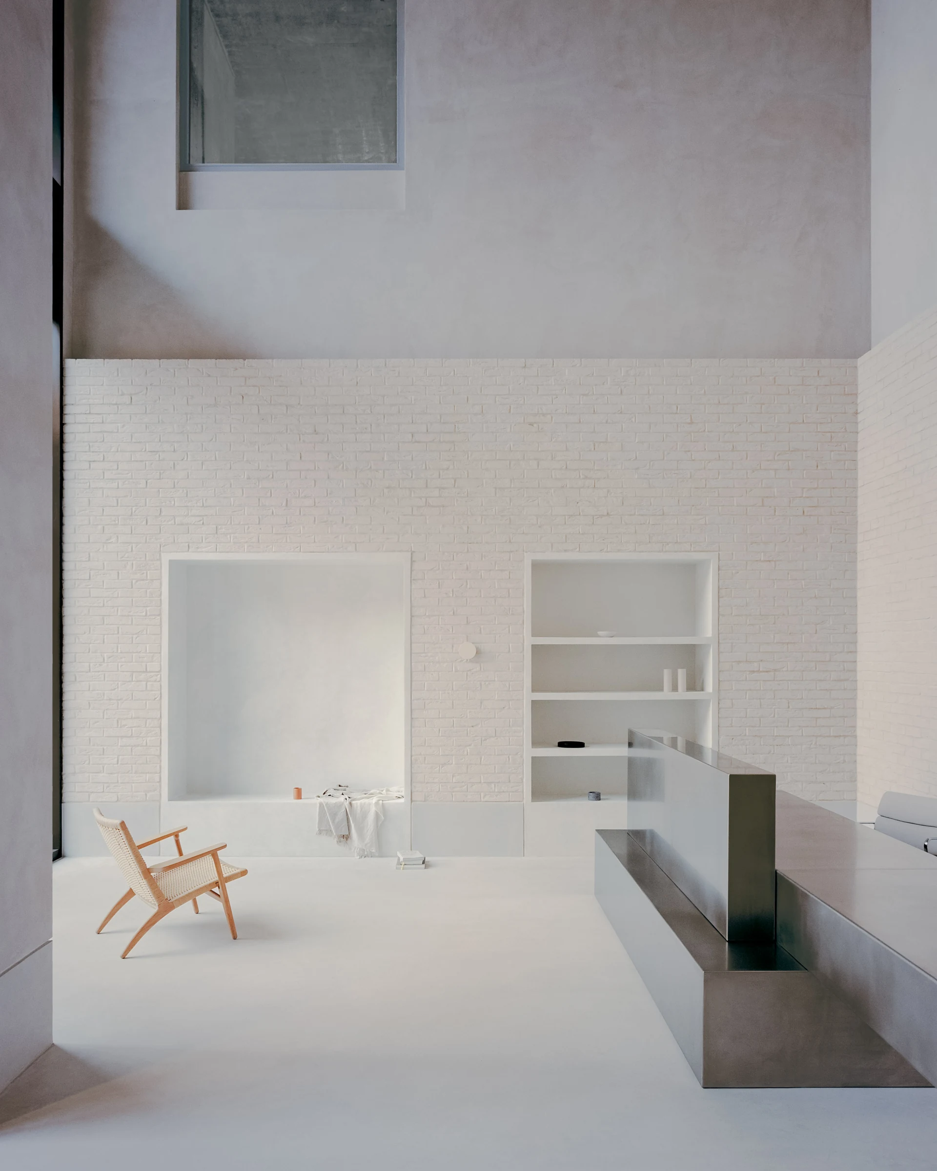 Cherry Orchard by Actual Office, photographed by Nick Dearden — Long view across the triple-height entrance hall: white-painted brick wall, pale poured concrete floor, the stainless steel concierge desk seen end-on with a Carl Hansen lounge chair at…