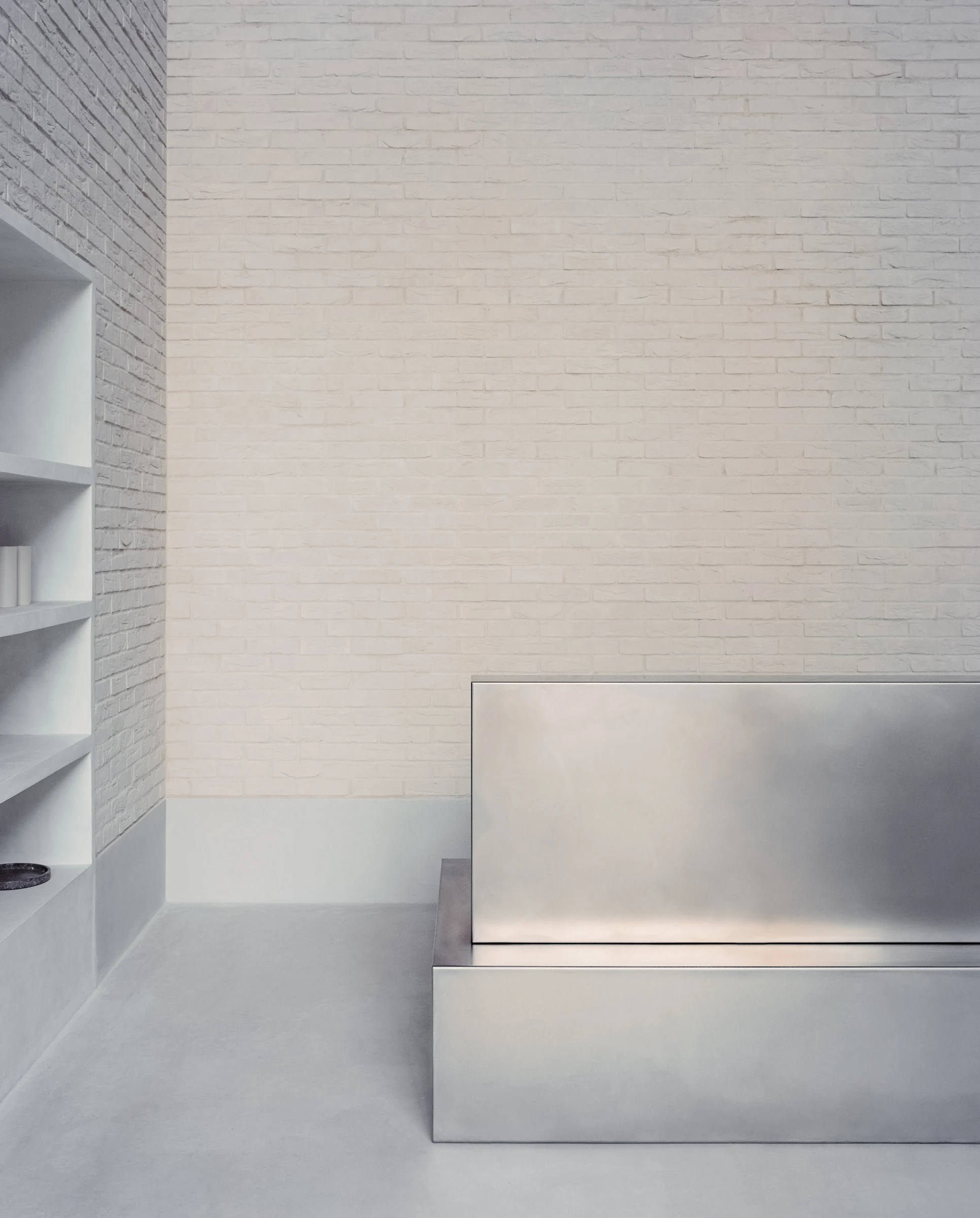 Cherry Orchard by Actual Office, photographed by Nick Dearden — Detail of the stainless steel concierge desk at the foot of the brick wall: brushed planes catching the room's pinkish wash, a recessed inset on the right side of the desk and a pale…