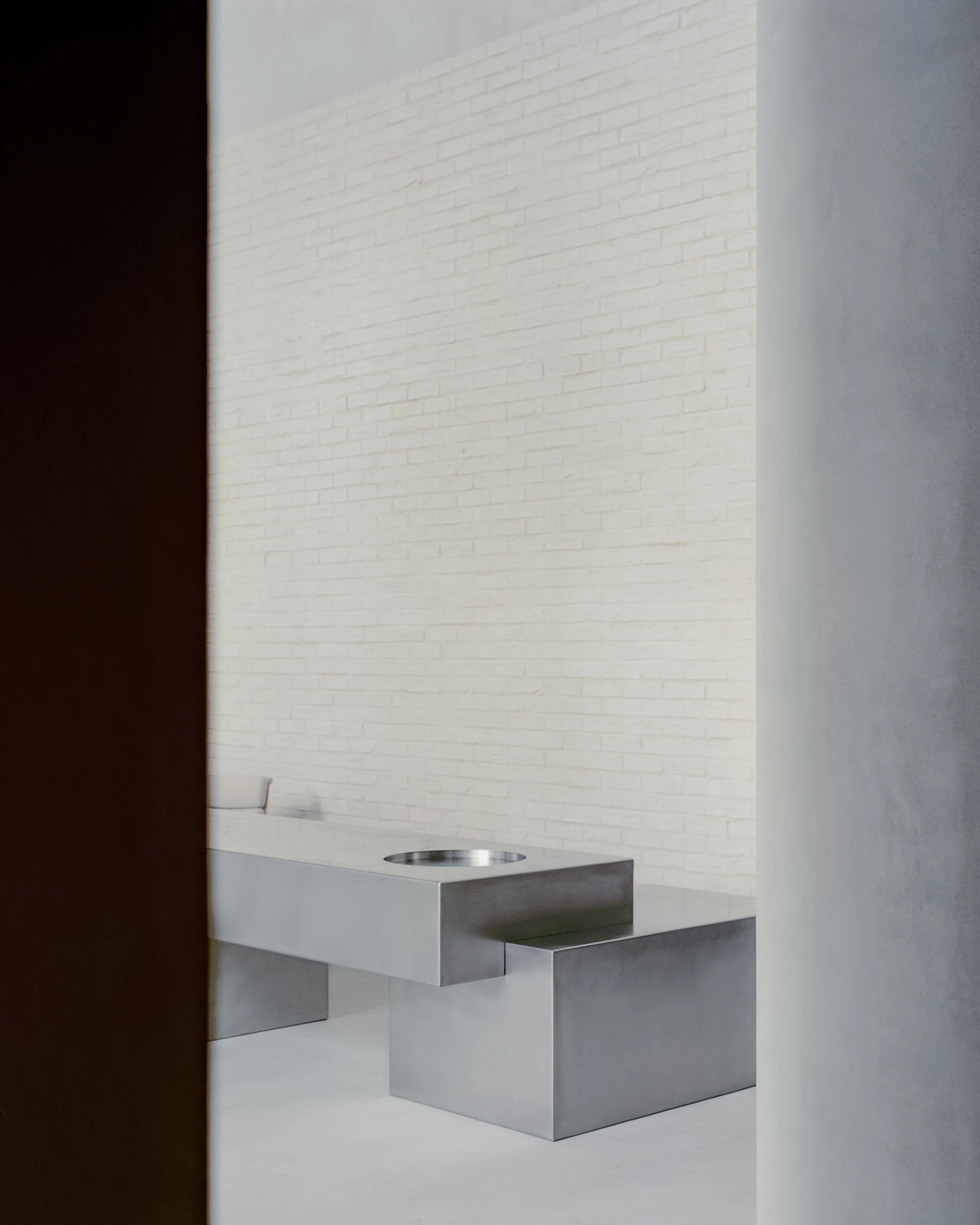 Cherry Orchard by Actual Office, photographed by Nick Dearden — The stainless desk seen through the dark slot of the cocoa-coloured chamber: deep brown wall at left, plastered column at right, the brushed-metal volume floating between the two against…