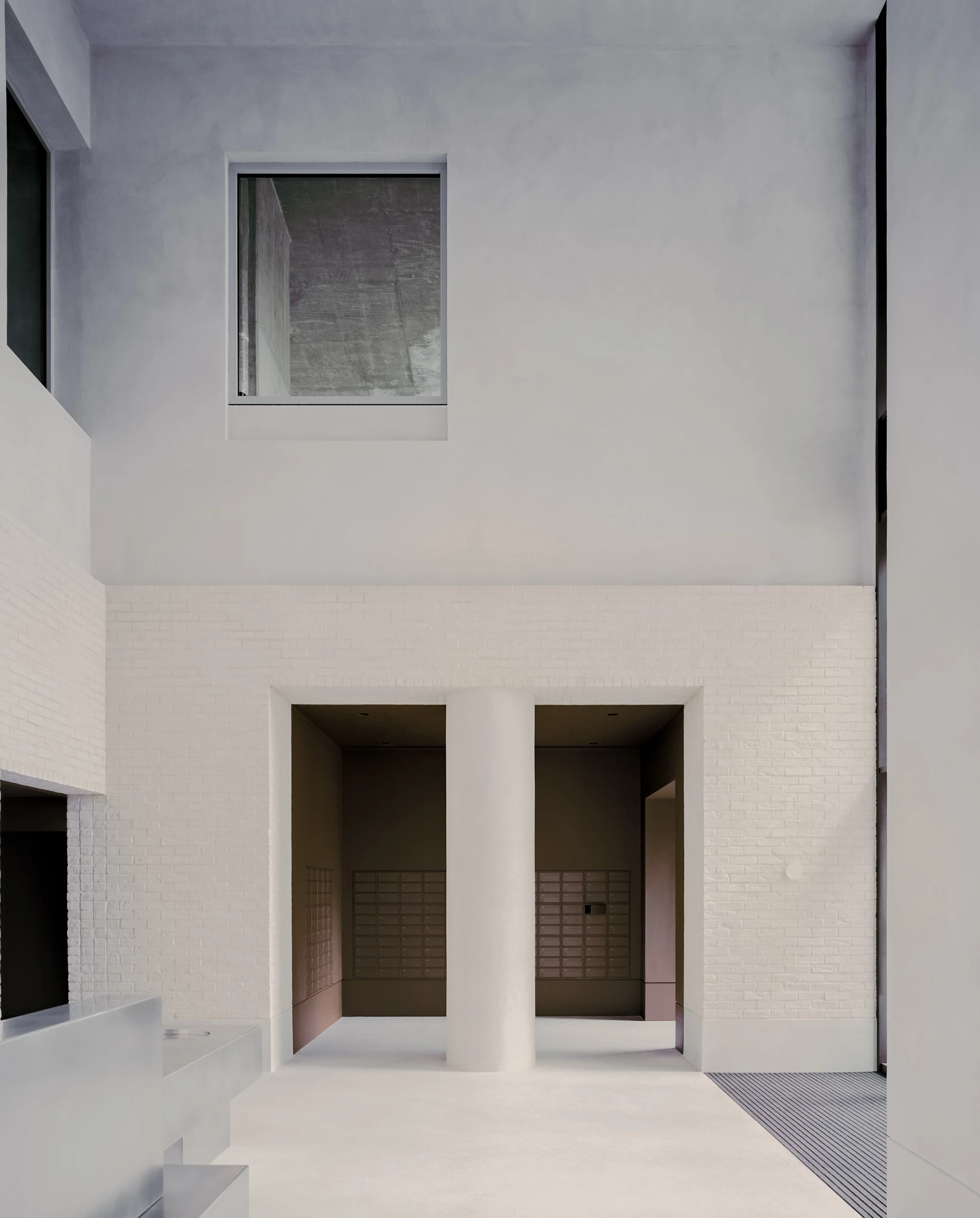Cherry Orchard by Actual Office, photographed by Nick Dearden — Frontal view of the entrance hall: a solitary plastered column frames the threshold to a deep cocoa-brown chamber holding two banks of stainless mailboxes, the concrete floor pale and the…