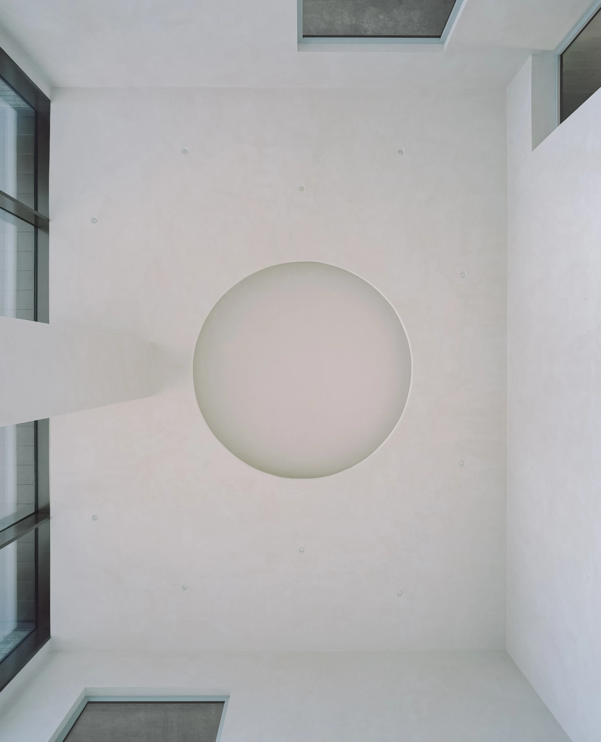 Cherry Orchard by Actual Office, photographed by Nick Dearden — Looking straight up through the triple-height void: a circular oculus cut into the pale plaster soffit, framed by smaller rectangular reveals at the edges, raking daylight from the side…