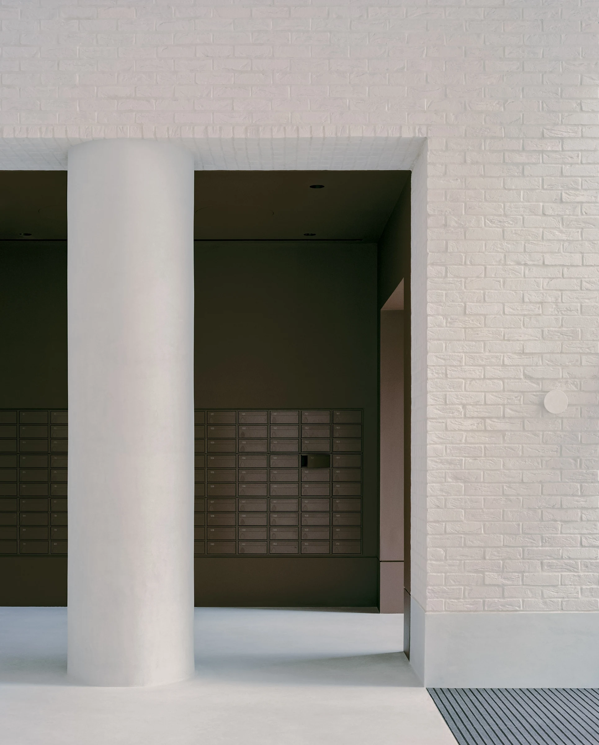 Cherry Orchard by Actual Office, photographed by Nick Dearden — The cocoa-pigmented mailbox chamber with a single Carl Hansen wishbone chair against the rear wall: stainless letterbox grid, plastered column at left, pale concrete floor across the…